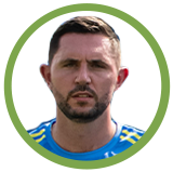 Close-up of a man with short dark hair and a beard, wearing a blue sports jersey, against a white background with a green circular border.