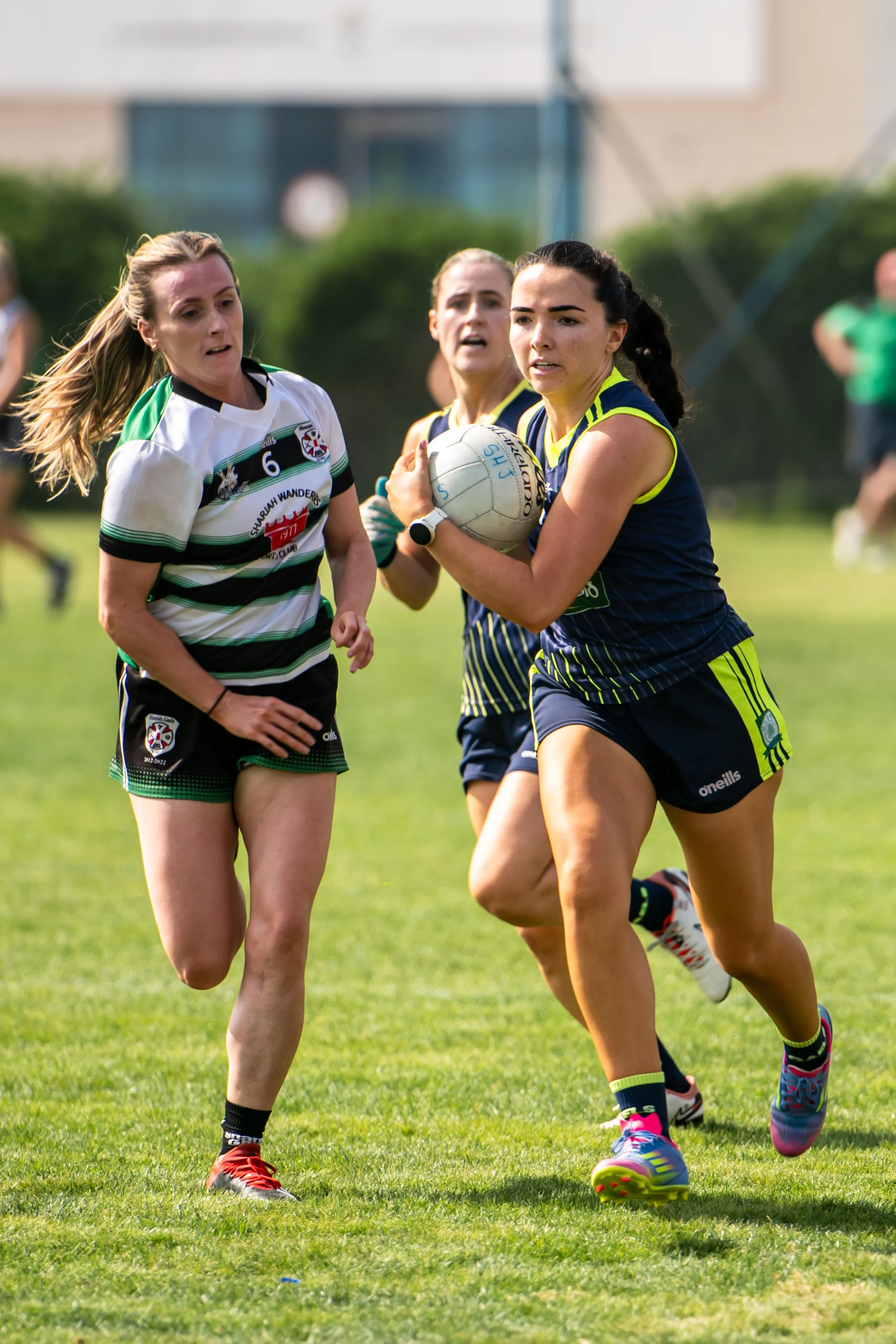 Women playing gaelic football on a grassy field, with one woman holding a gaelic ball and others running beside her, all wearing team uniforms and colorful athletic shoes.