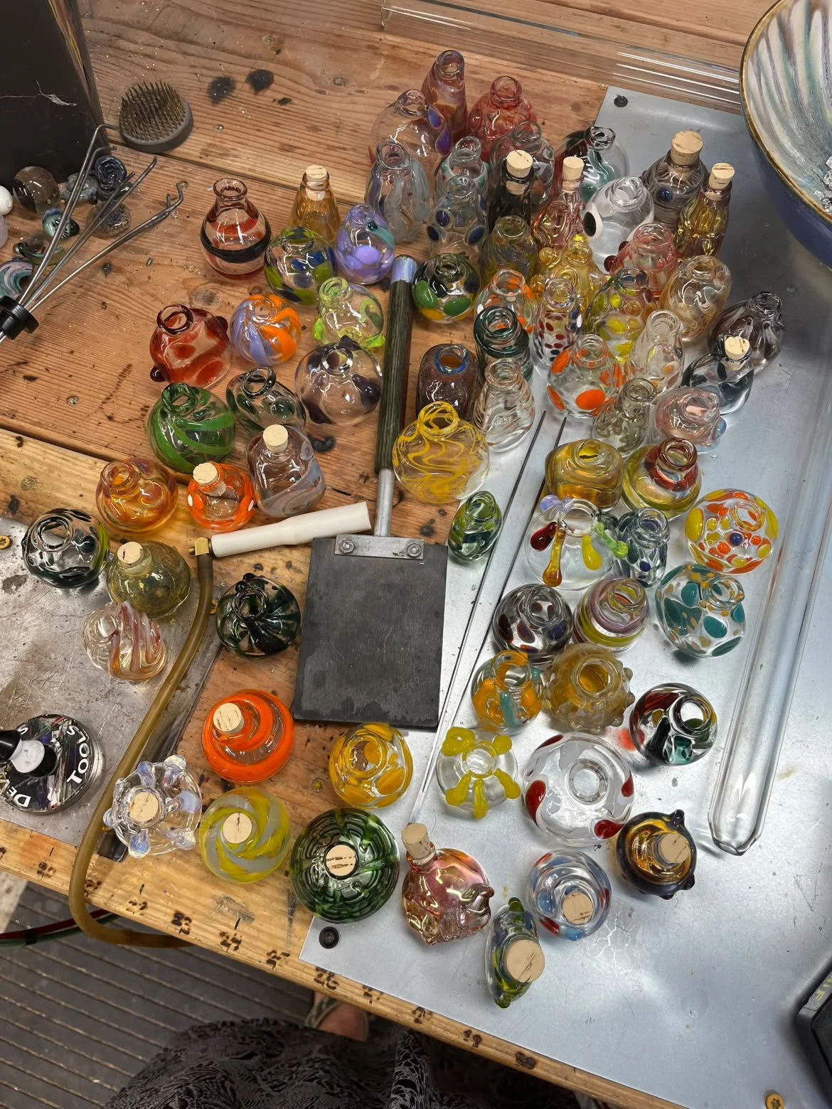 Colorful glass art pieces being crafted on a workspace. Various glass objects in different shapes, sizes, and colors are spread across a wooden and metal surface, with some tools and tools parts visible.