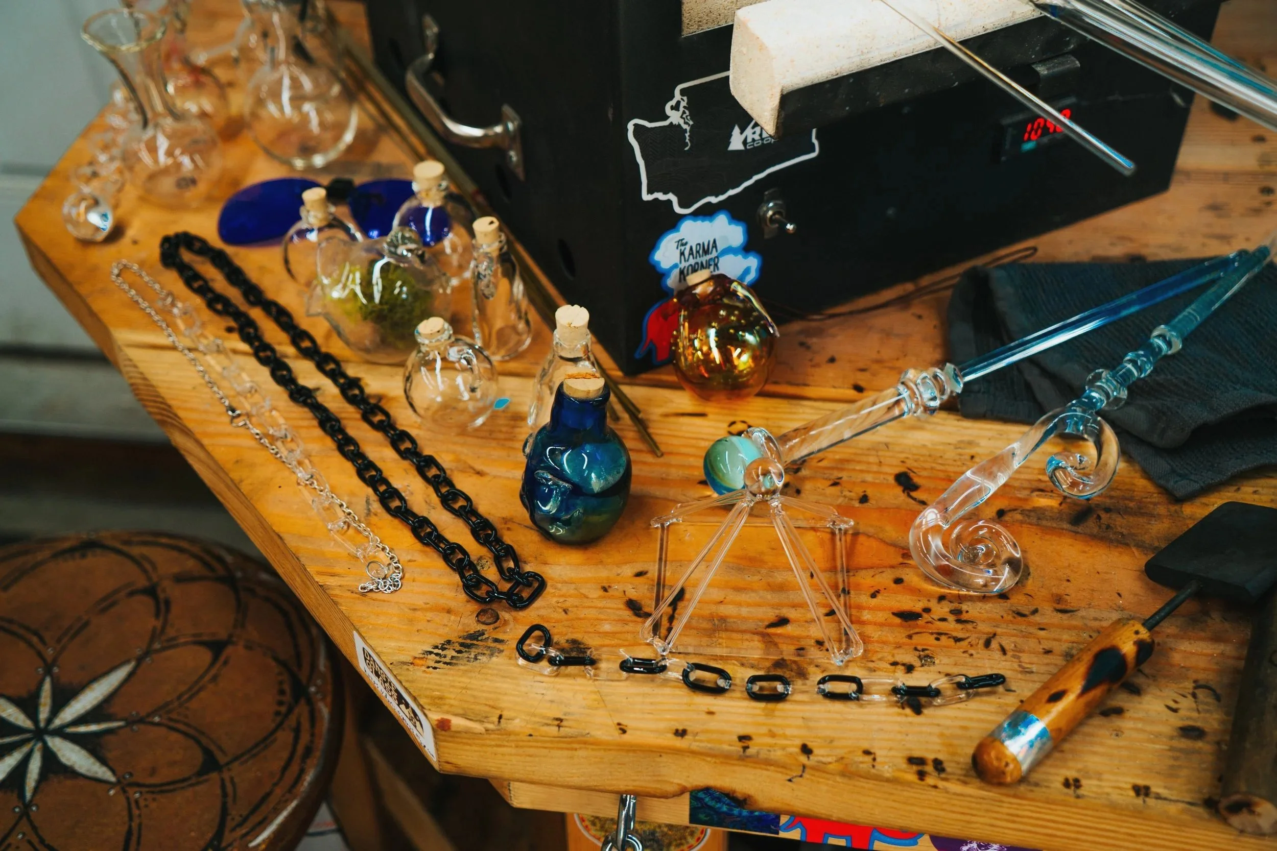 Display of various unique hand made glass pieces, including necklaces, pendants, and decorative glass objects, arranged on a wooden table.