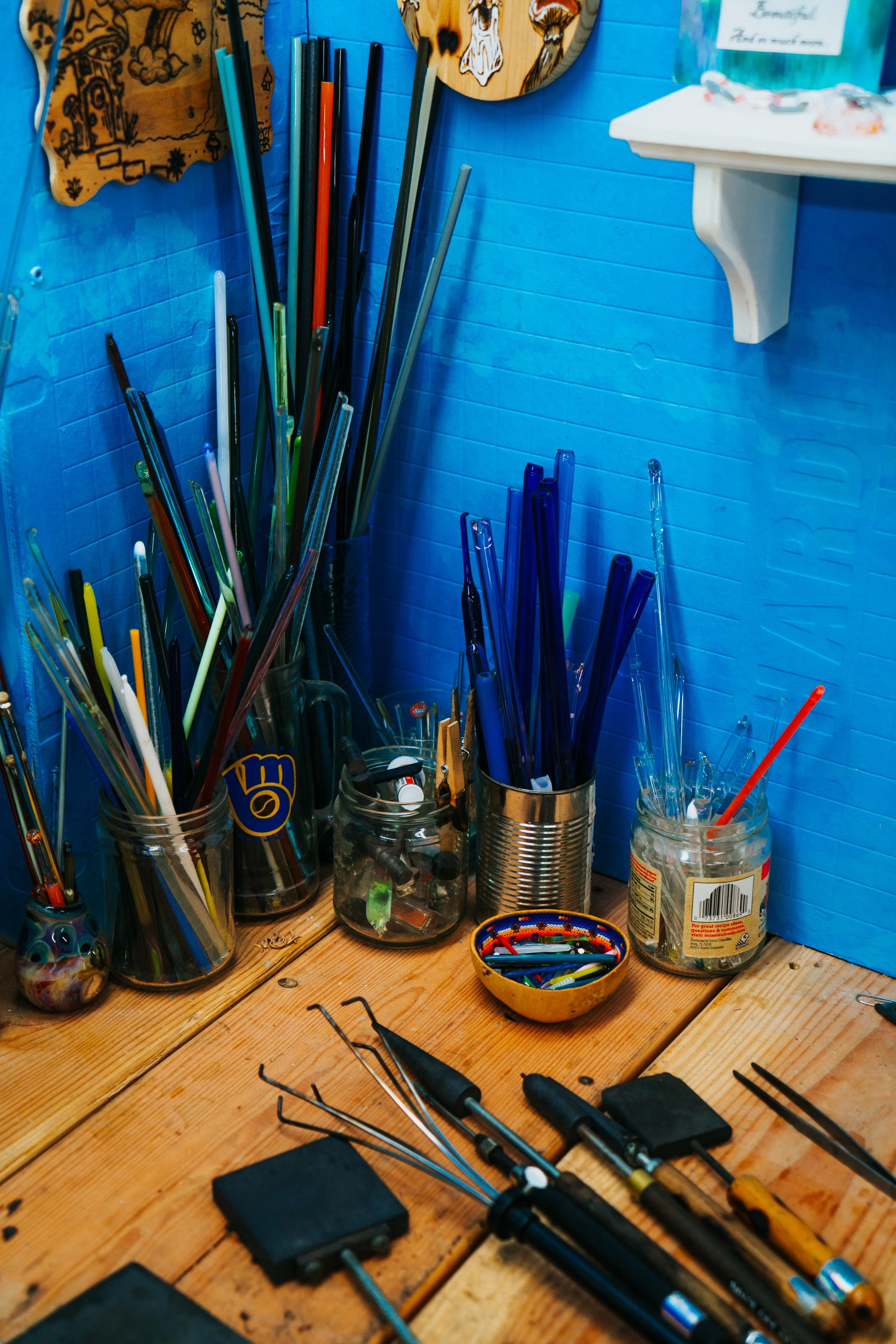 A collection of glass jars filled with colorful glass blowing materials. Various glass blowing tools and equipment are scattered across the table, including metal tools and a small bowl of multicolored beads.