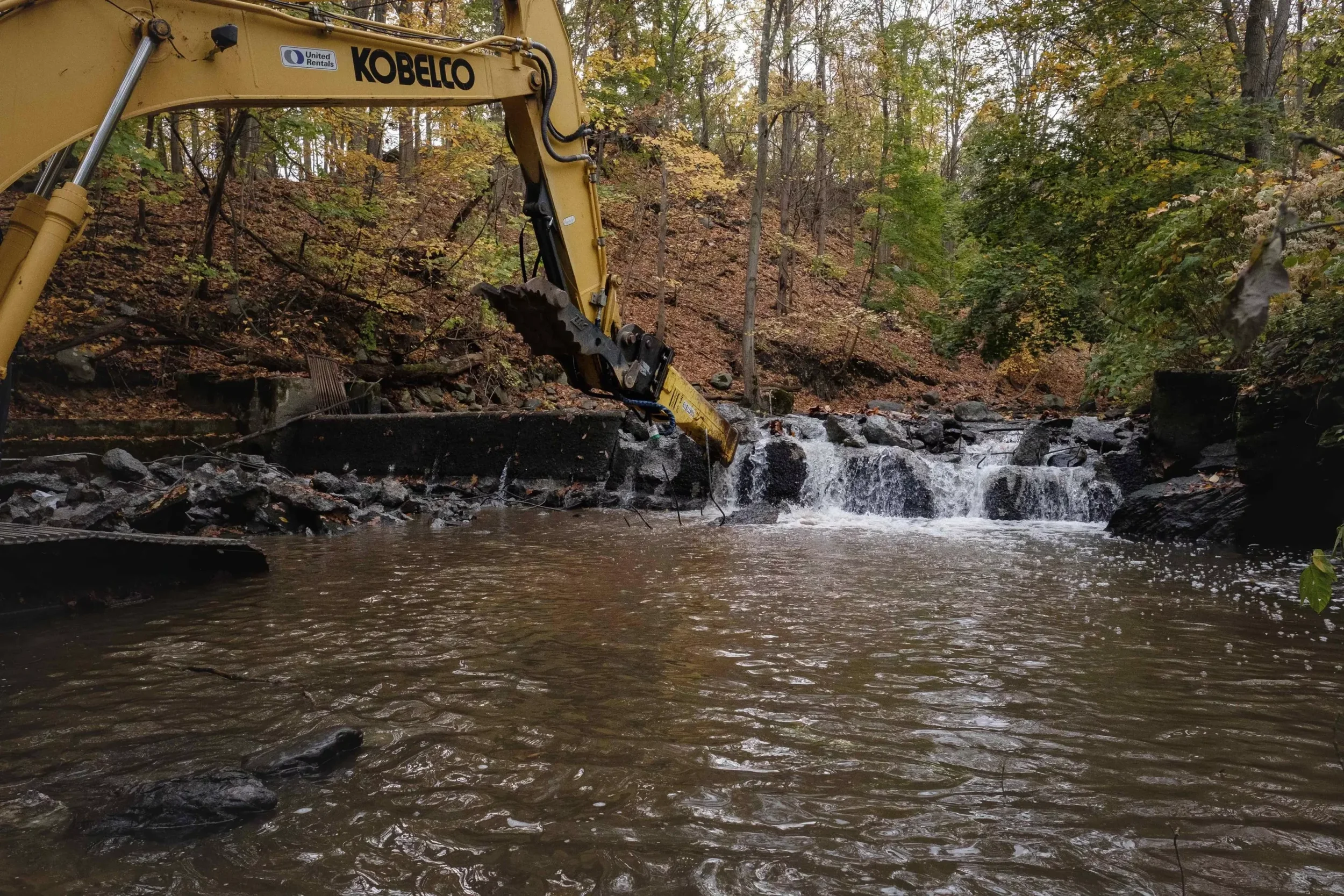 A large yellow construction excavator working on a creek with water flowing over rocks, surrounded by a forest with autumn-colored trees.