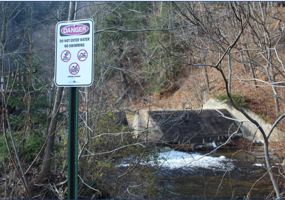 A warning sign near a river indicating danger, no swimming, no entry into water, and a strong current, with a small waterfall and leafless trees in the background.