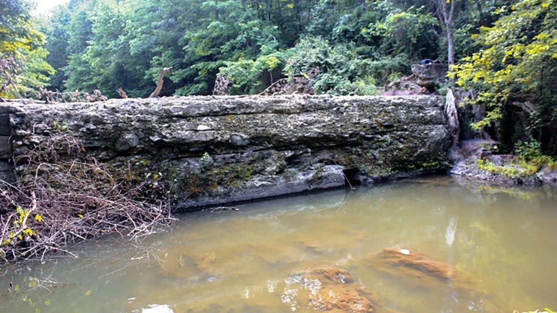 A large, moss-covered rock formation over a shallow water stream in a lush, green forest.