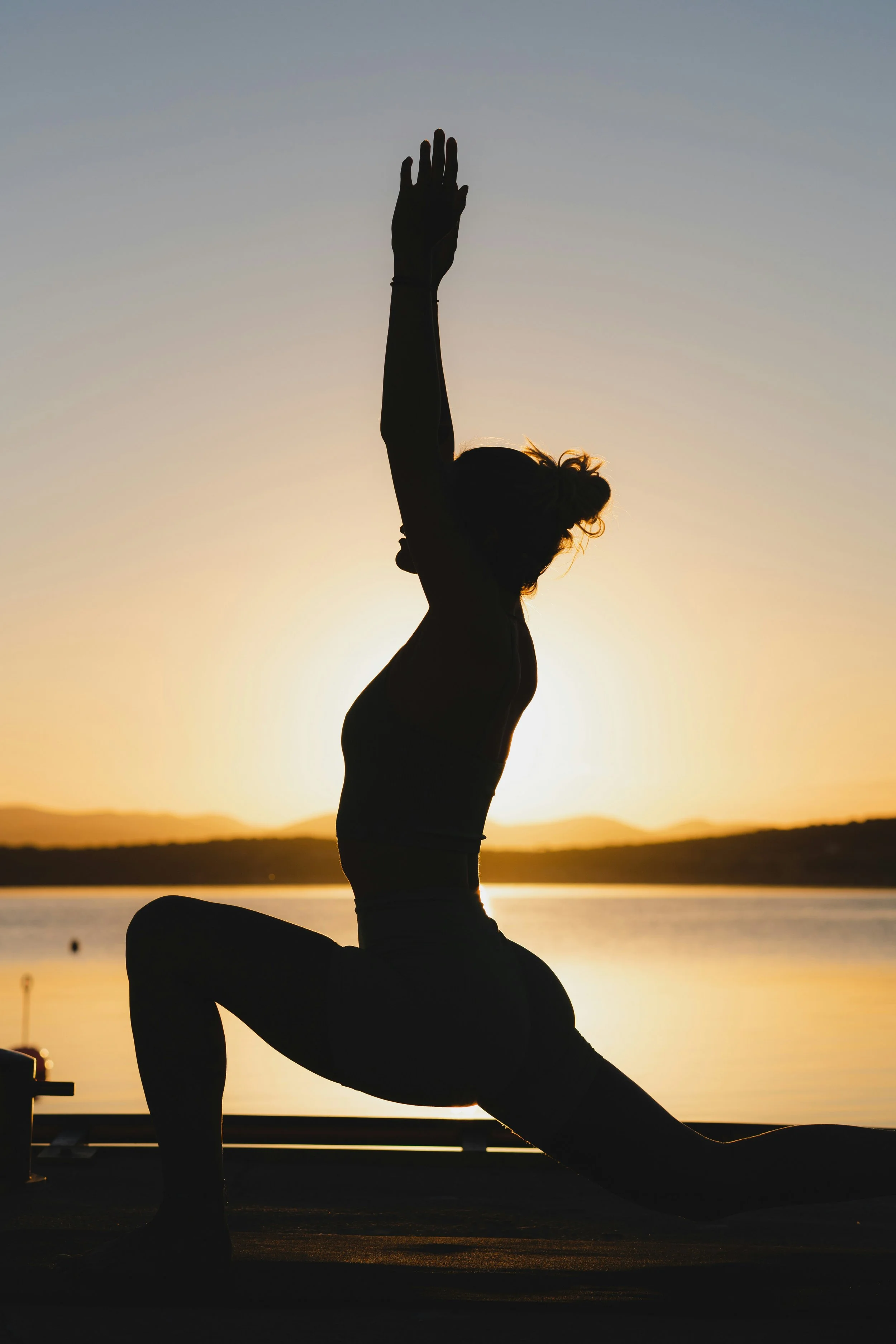 Silhouetted person practicing yoga or stretching at sunset near a body of water, with mountains in the background.