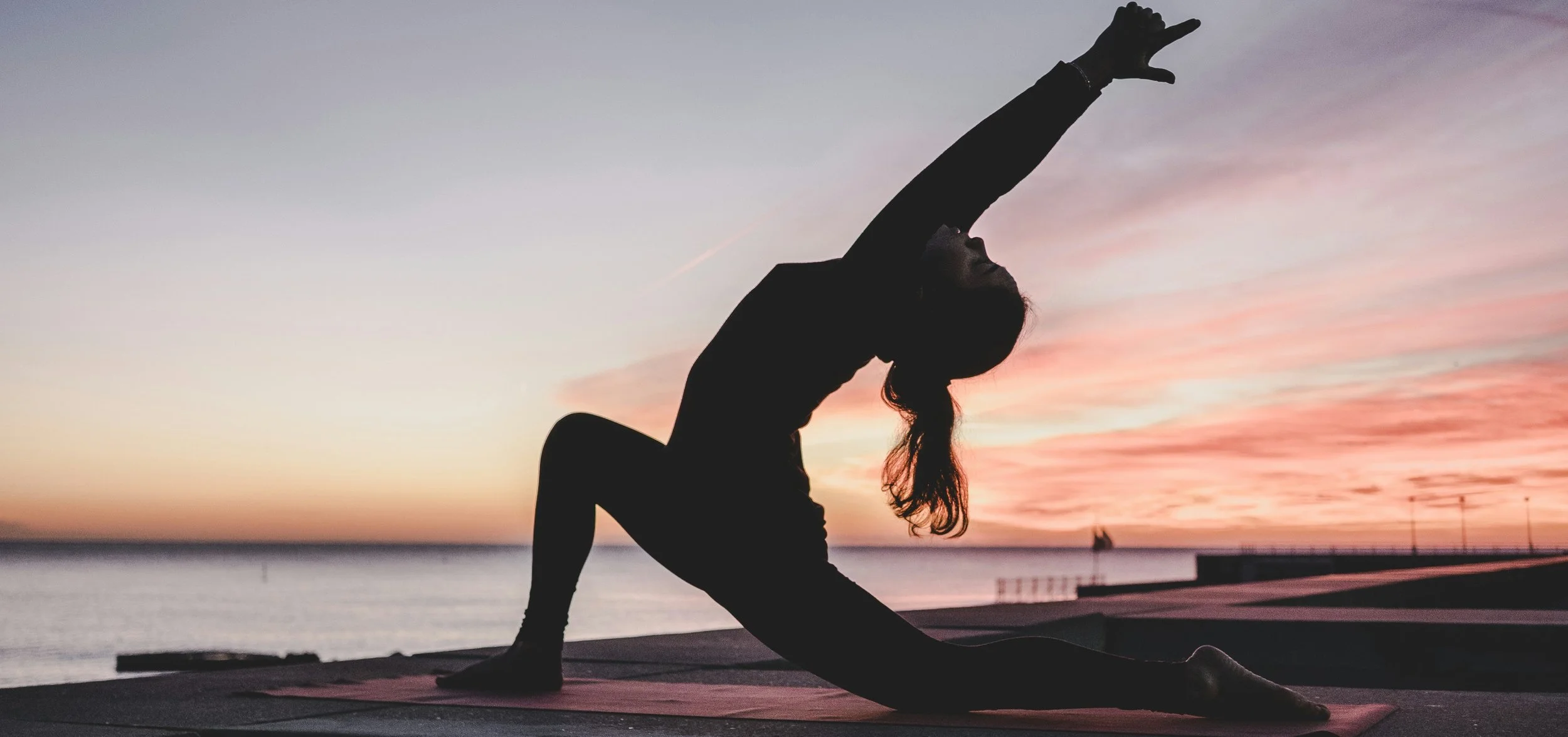 Silhouette of a woman practicing yoga on a mat by the water during sunset, performing a side stretch with one arm reaching upward.