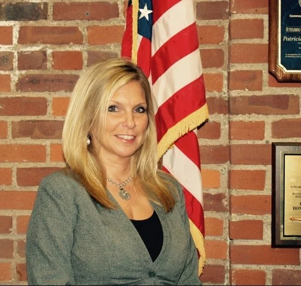 A blonde woman in a gray blazer stands indoors next to an American flag and a brick wall with framed documents.