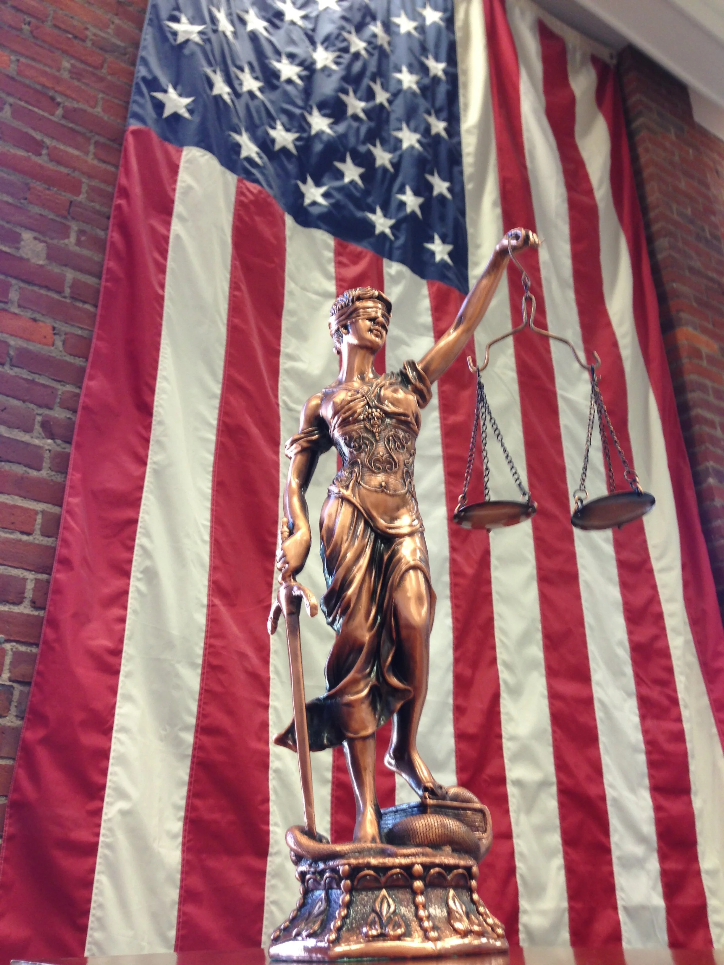 A bronze statue of Lady Justice with a blindfold, holding scales in her left hand and a sword in her right hand, in front of an American flag.