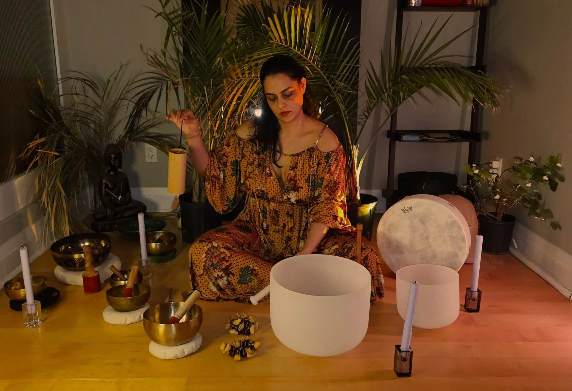 A woman sitting cross-legged on the floor in a dimly lit room, holding a yellow chime, surrounded by various musical instruments, candles, and plants, with a shelf and a drum in the background.