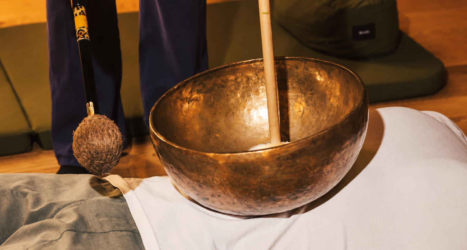 A large, bronze singing bowl with a wooden striker inside, placed on a white cloth on a table.