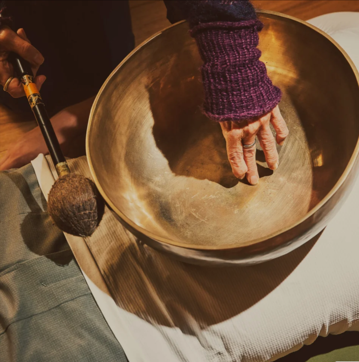 A person with a purple sweater playing a singing bowl with their hand, while another person holds a mallet nearby.
