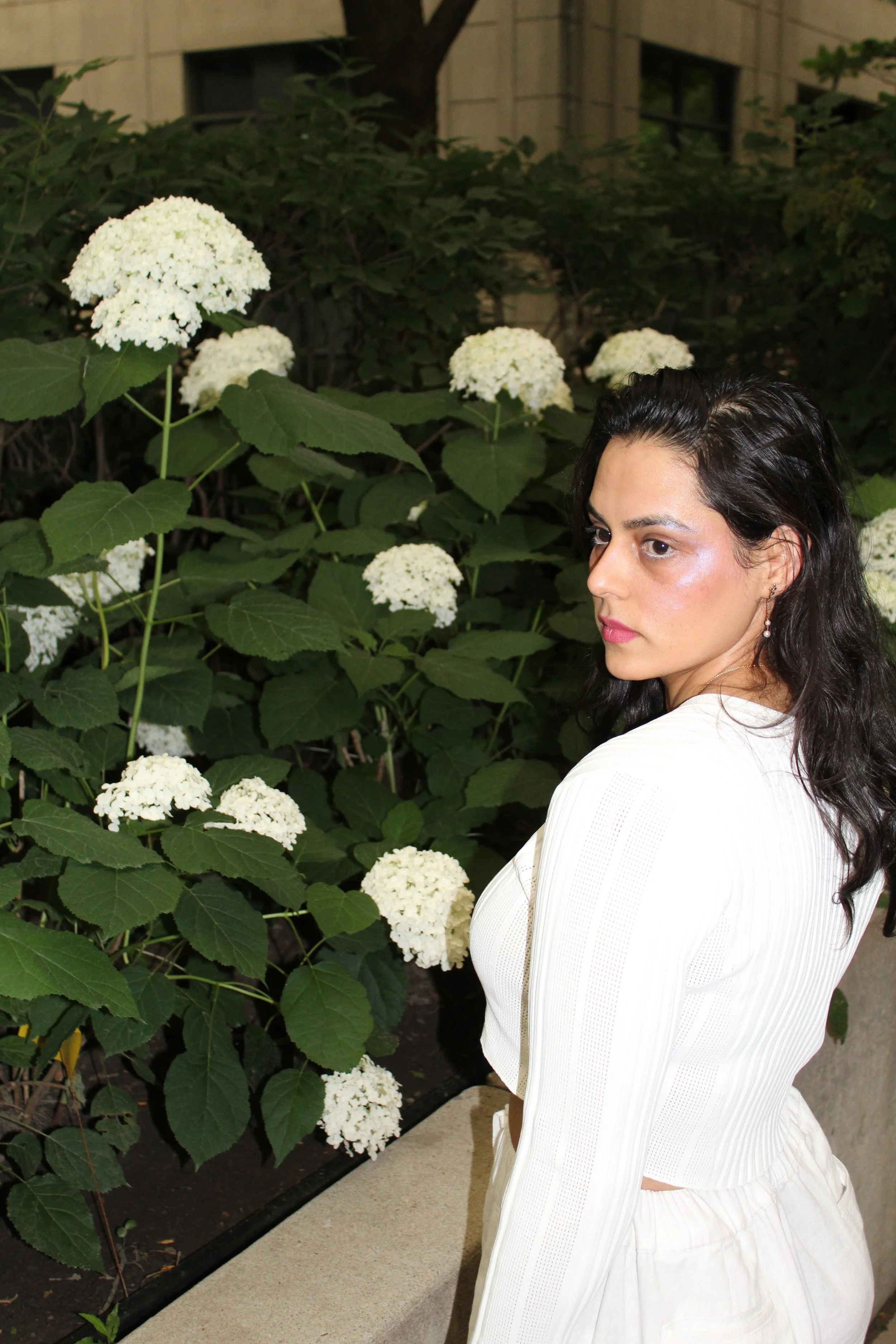 A woman with dark hair and earrings standing outdoors near white flowers and green leaves.