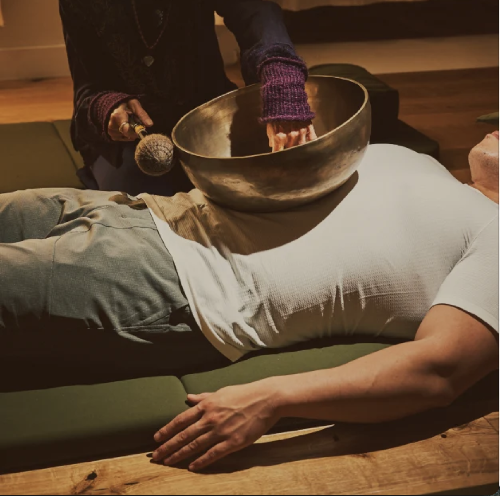 Person lying on a green massage table with their arm resting on the edge, while another person is holding a singing bowl and a mallet. The setting appears to be a calming room, possibly during a sound healing or meditation session.