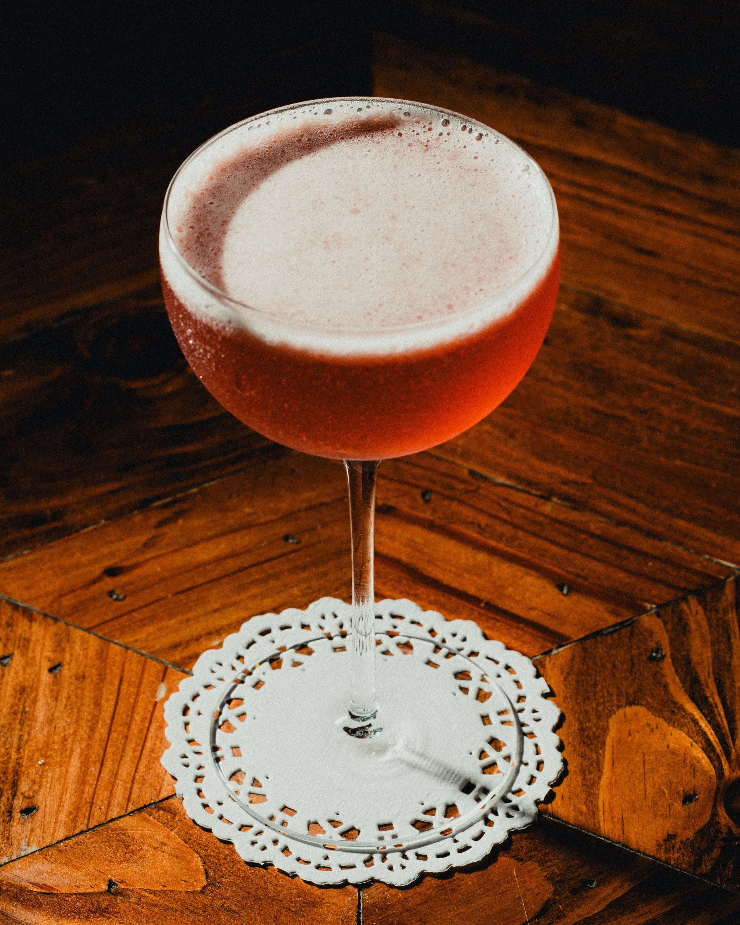 A glass of rosé wine with a frothy head on a wooden table, resting on a decorative white paper doily.