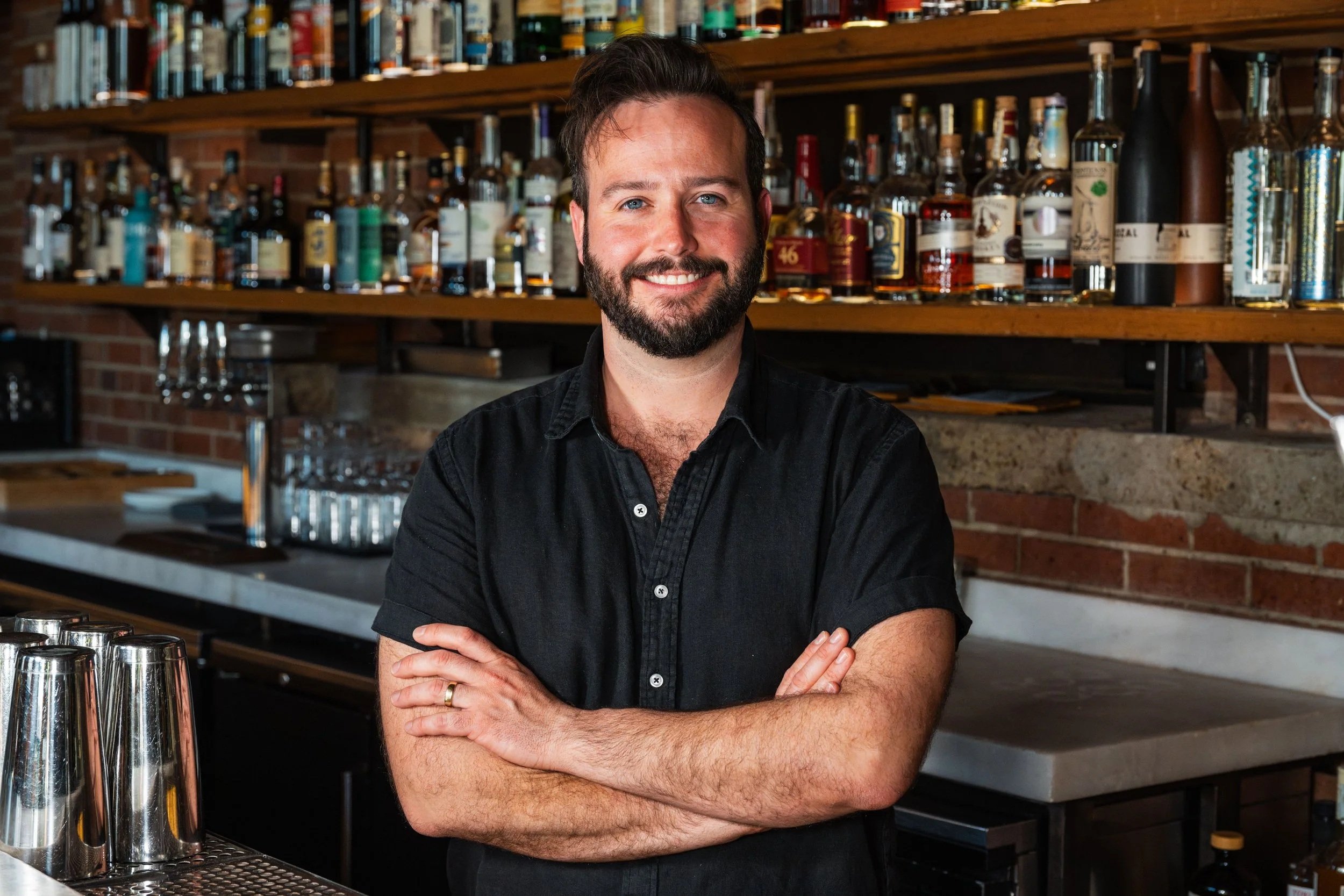 A smiling man with a beard and blue eyes standing with arms crossed in a bar or pub, with shelves of liquor bottles in the background.