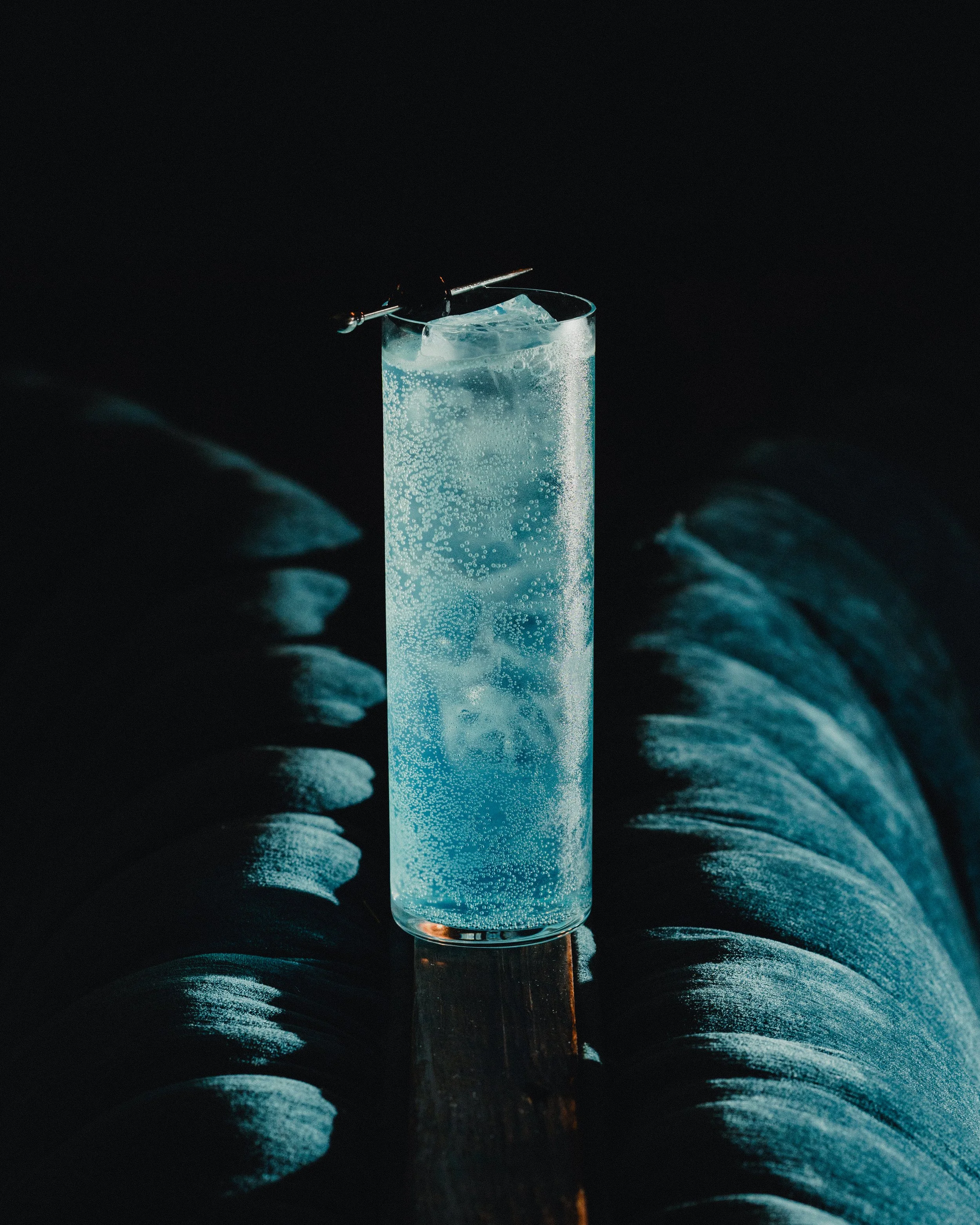 A tall glass of clear soda with ice cubes and bubbles, with a metal cocktail pick resting on the rim, placed on a dark surface with a reflective sheen, and a dark background.