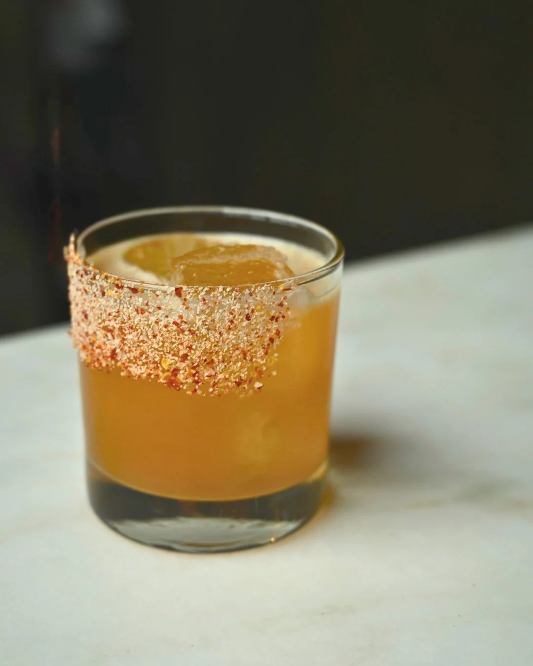A glass of yellow-orange cocktail with a salt and chili rim, served on a white surface, with a dark background.