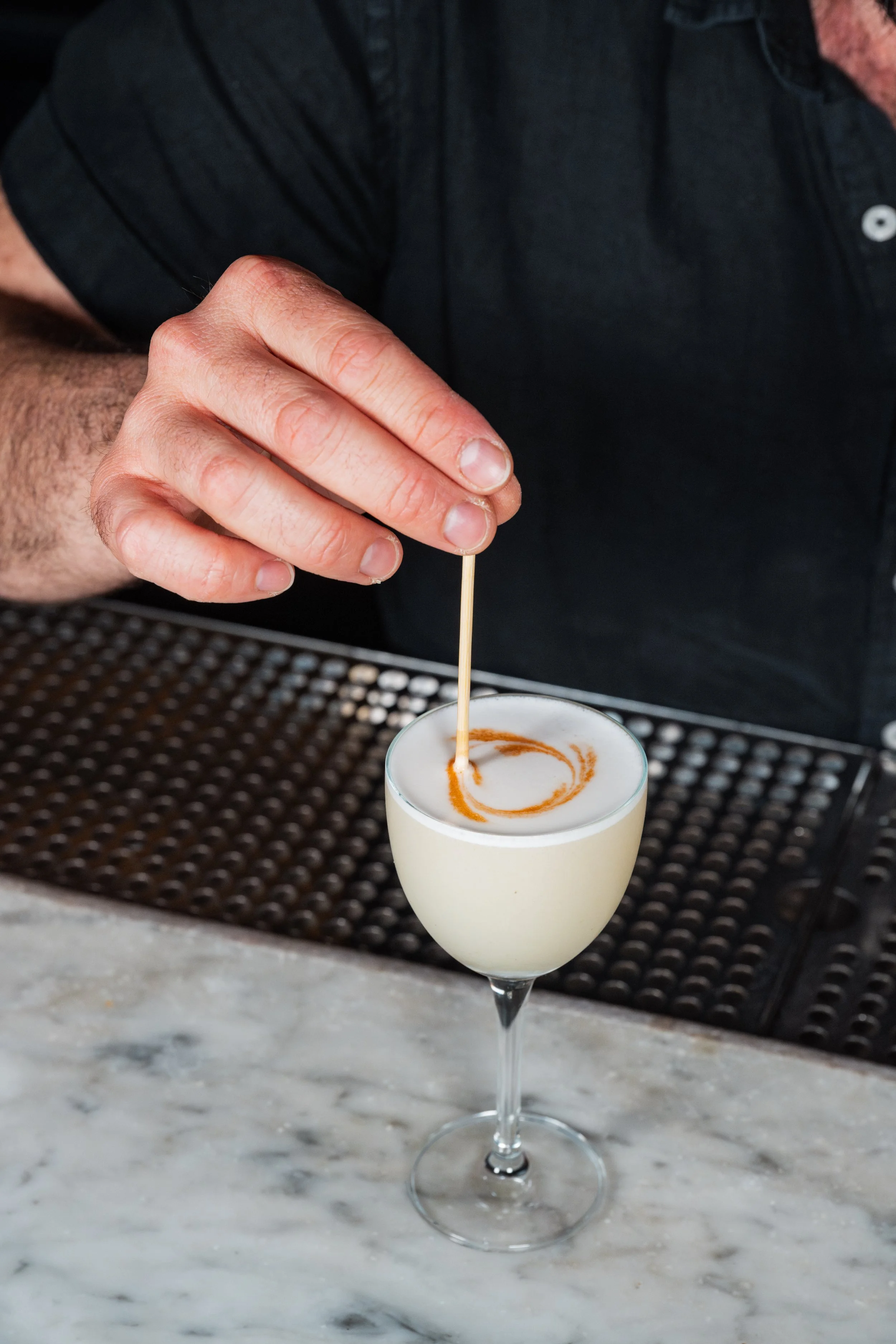 A person in a black shirt is making a cocktail by adding a stir stick to a frothy, creamy drink garnished with a drizzle of caramel or similar syrup, in a coupe glass on a marble countertop.