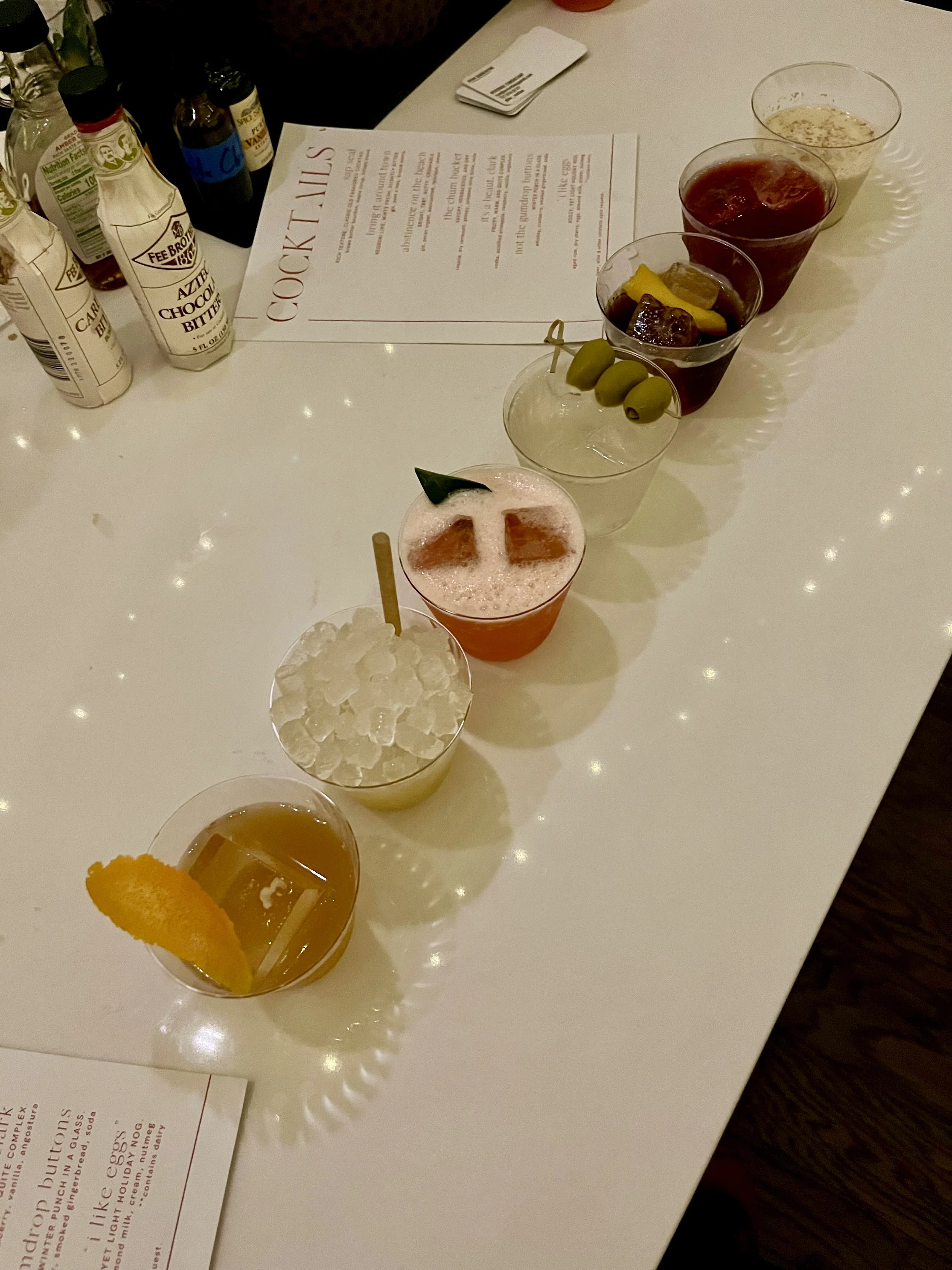 A table with cocktail drinks, a menu, and condiment bottles. The drinks include a pink cocktail with an orange slice and a cherry garnishment, a lemon-lime drink with green olives on a pick, and various other beverages. Condiments include Vicks VapoRub, allergy medicine, and a jar of syrup.