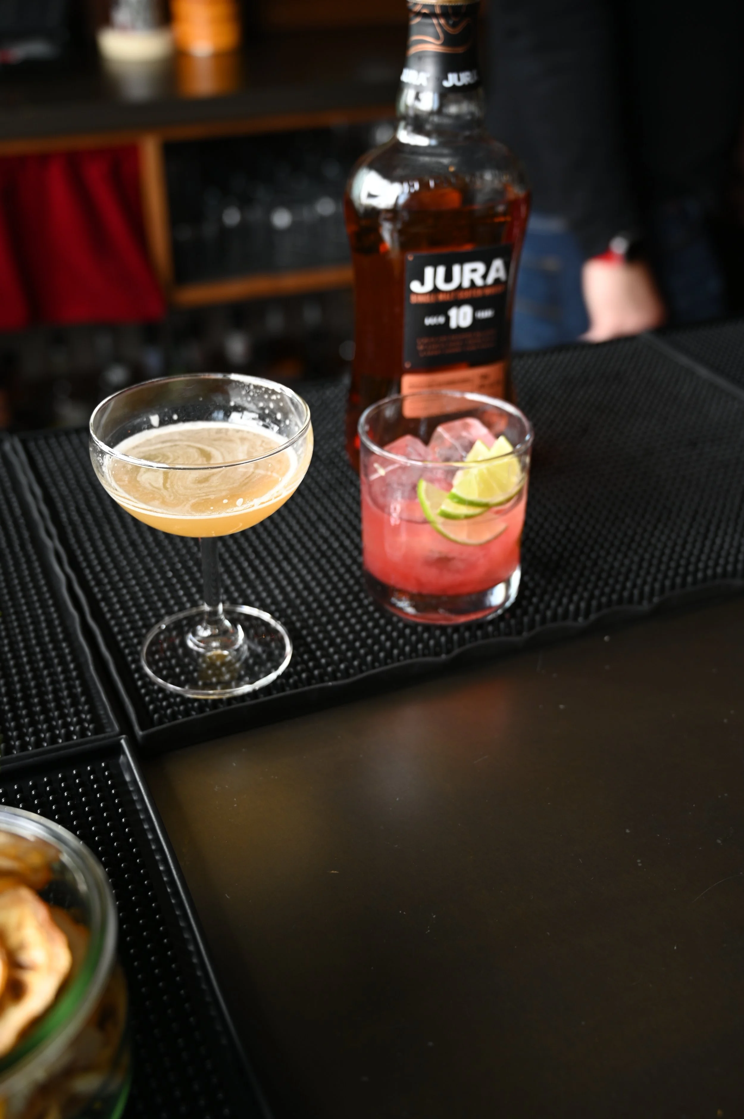 A bar scene with a bottle of Jura am 10 whiskey, a glass of pink cocktail with lime slices, and a cocktail glass with a light-colored mixed drink on a black rubber bar mat.