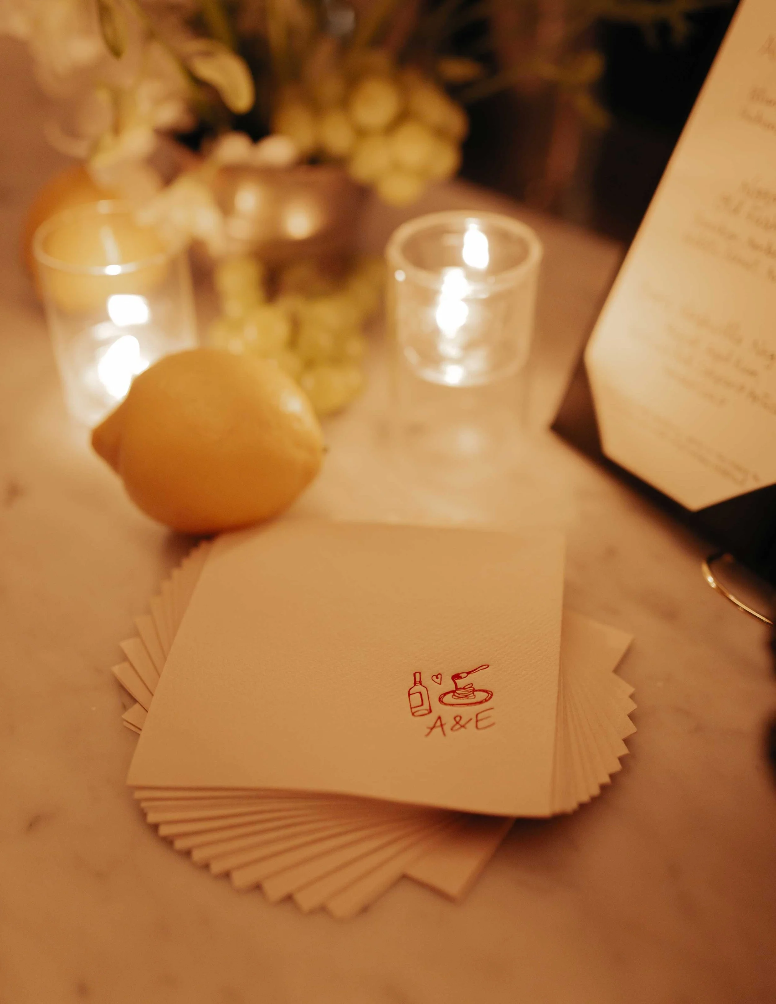 Close-up of a stack of cream-colored paper napkins with a red logo that includes a wine bottle, a wine glass, and a plate with a fork and knife, along with the text 'A & E'. In the background, there are two small candles in glass holders and a lemon in the foreground, with a blurred menu on the right side.