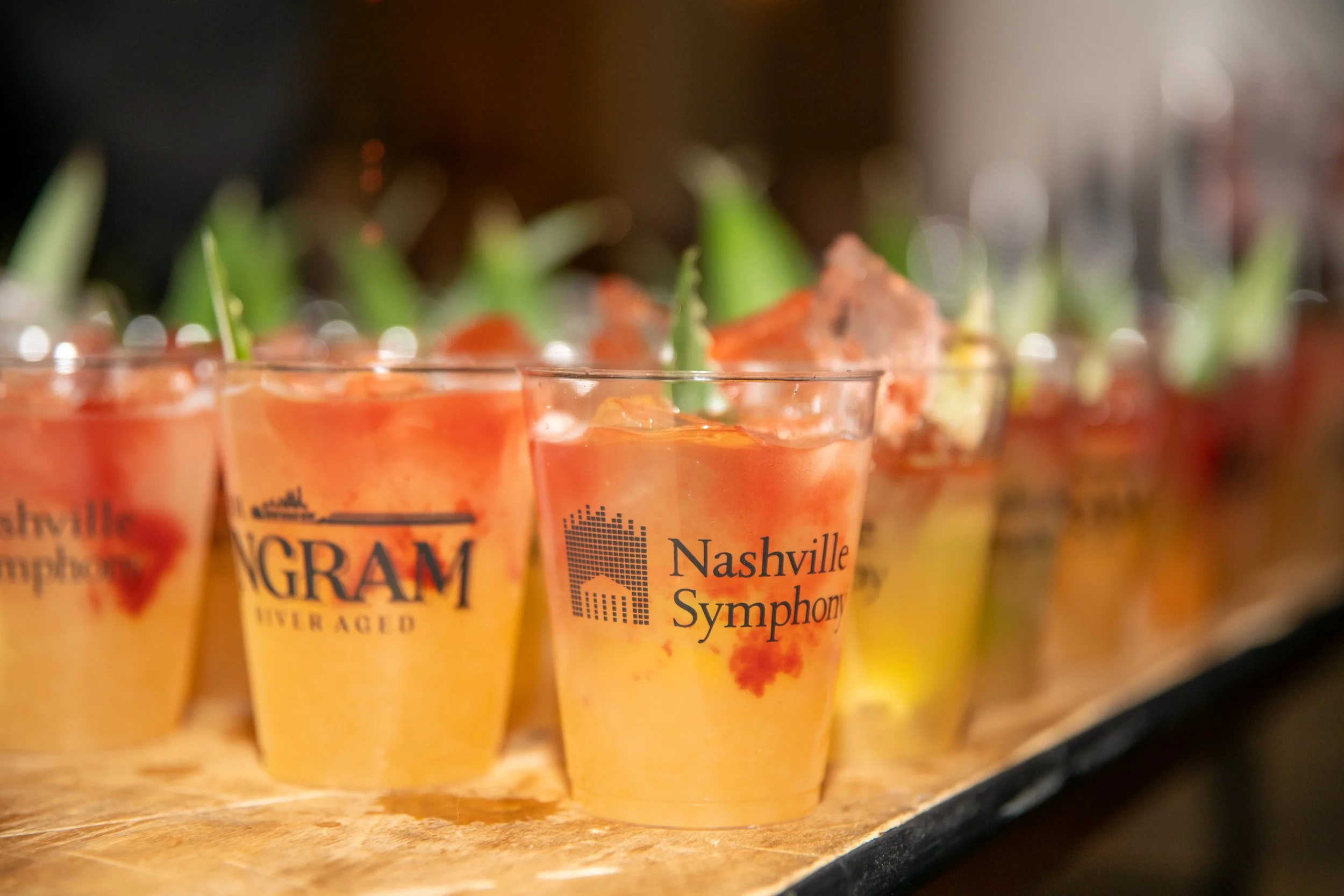 Close-up view of small plastic cups filled with a peach-colored beverage, labeled 'Nashville Symphony,' arranged on a wooden surface.
