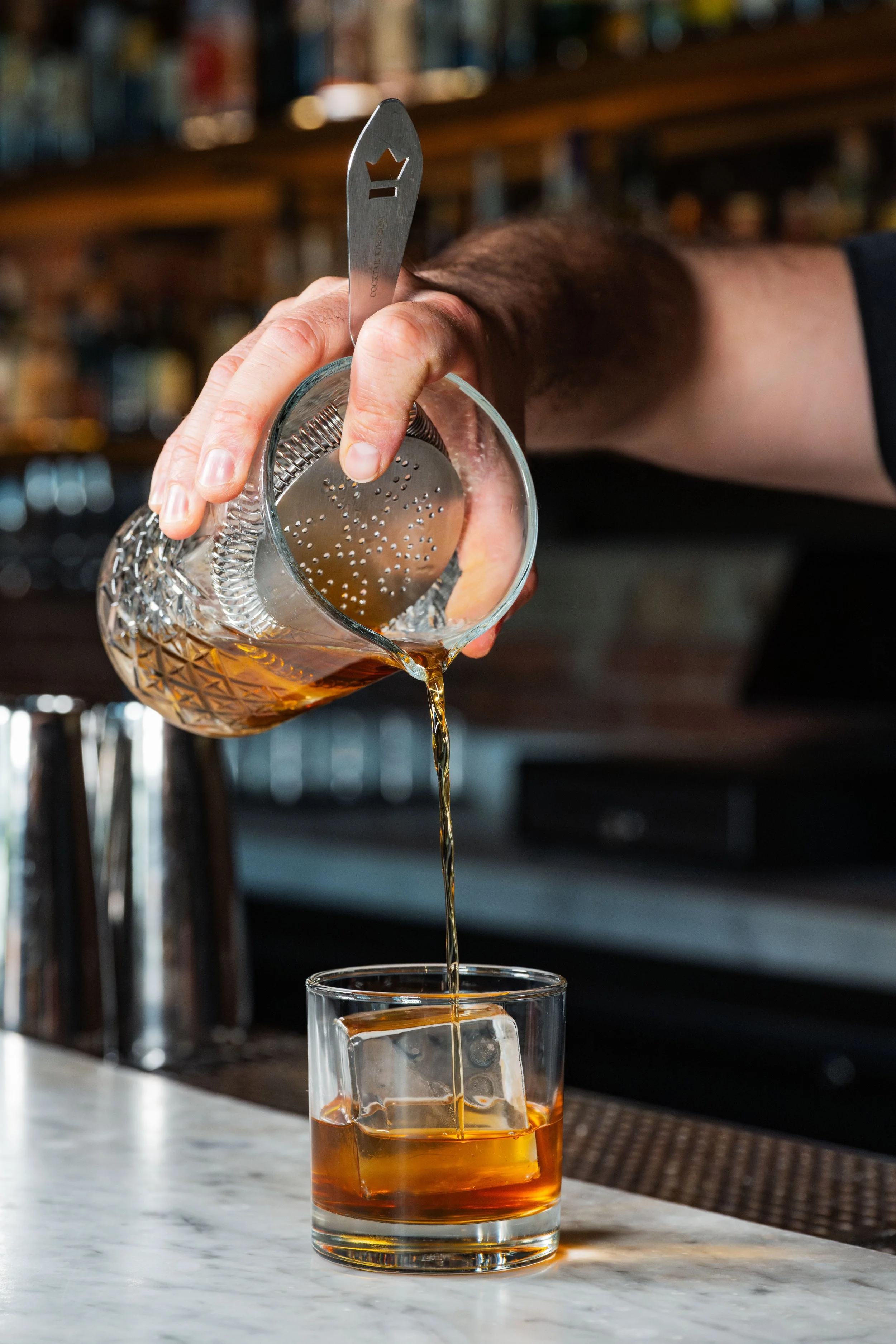 A hand pouring an amber-colored drink from a glass pitcher into a clear glass on a marble countertop, with a bar background.