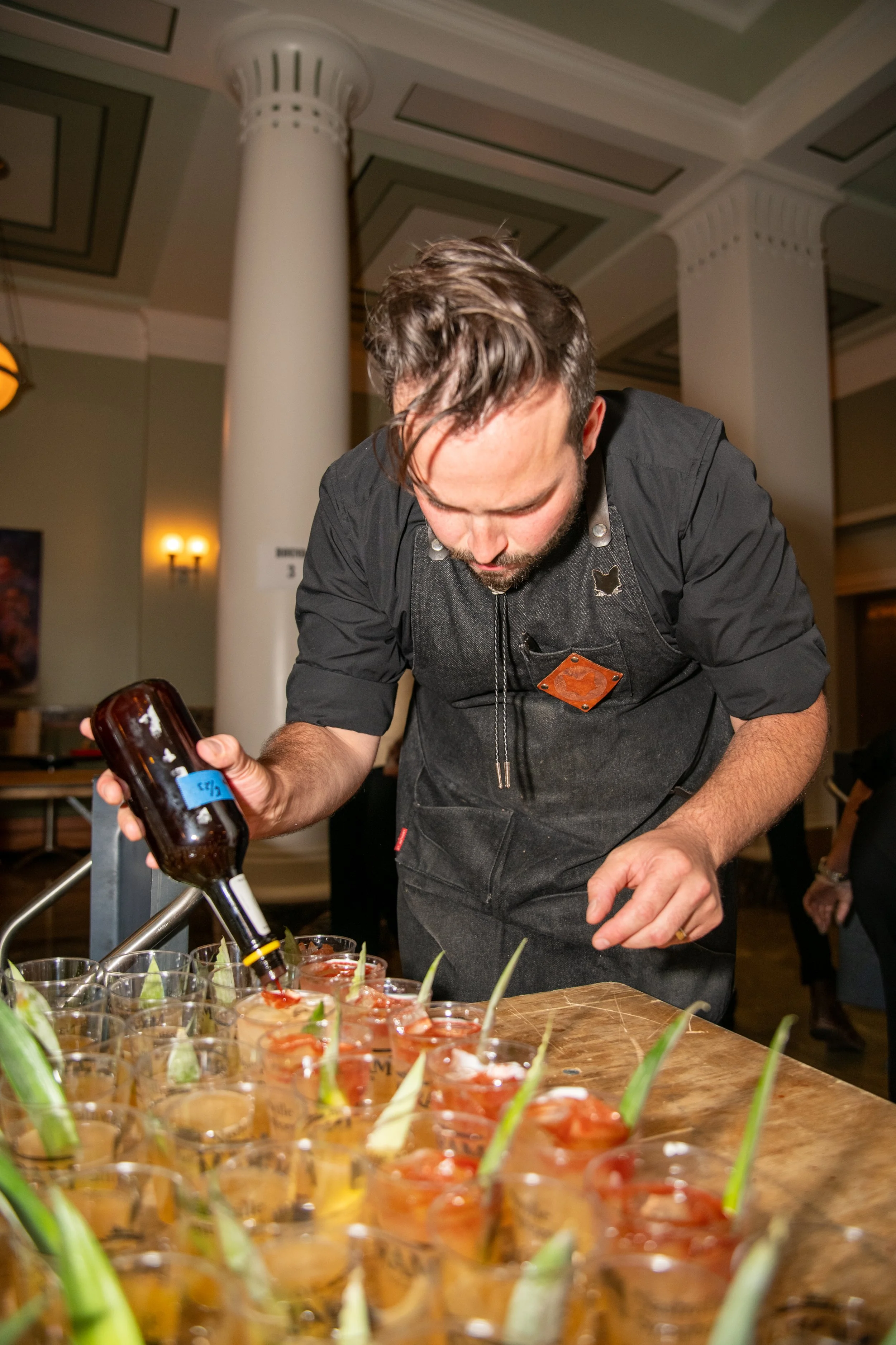 A man with styled brown hair and a beard, wearing a black shirt and apron, is pouring a drink into a glass at a table with many glasses filled with drinks garnished with pineapple and lime slices in an indoor setting.