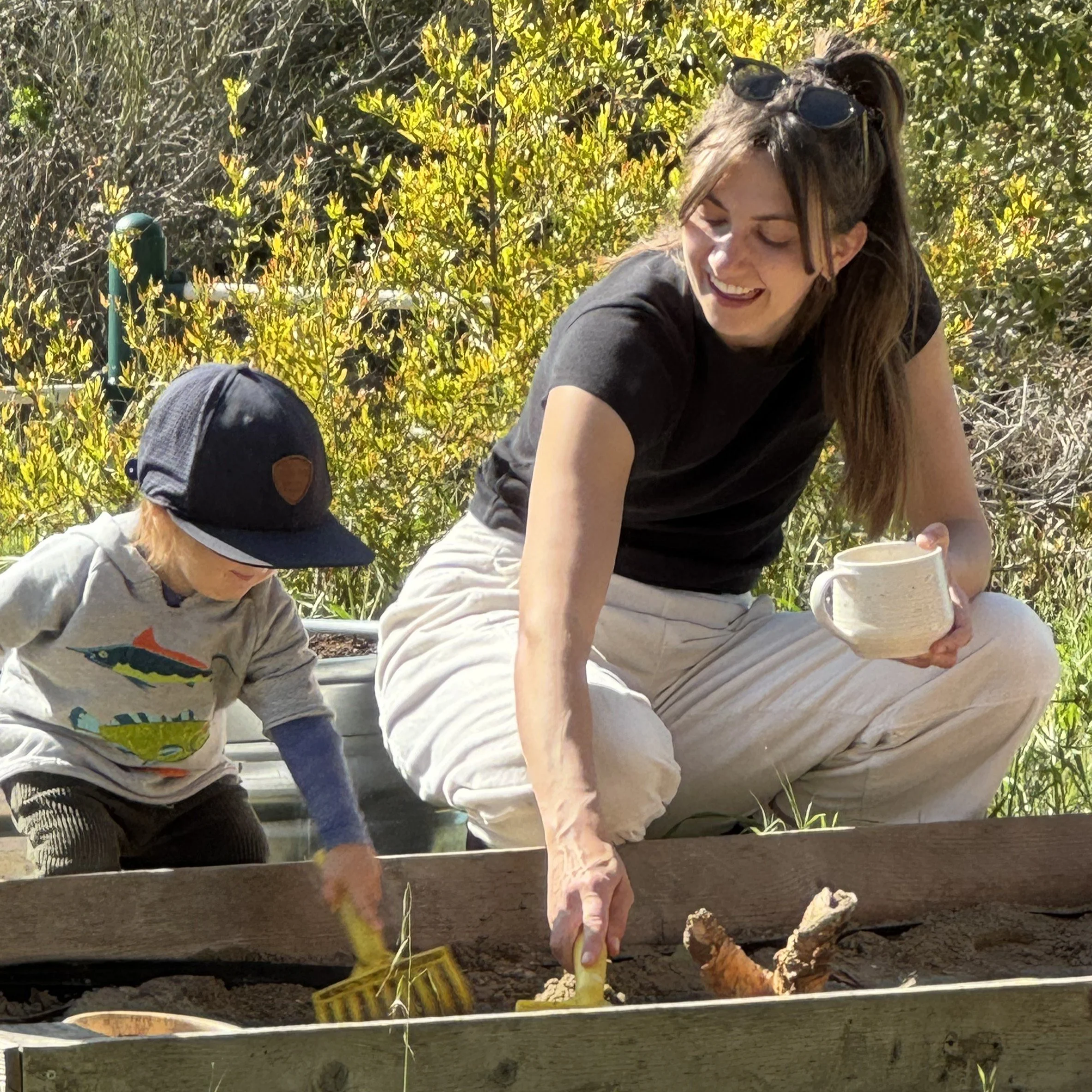 A woman and a young child gardening outdoors. The woman wears sunglasses on her head and a black shirt, smiling while working with soil. The child wears a baseball cap and a light-colored hoodie, focused on the soil, with a garden and foliage background.
