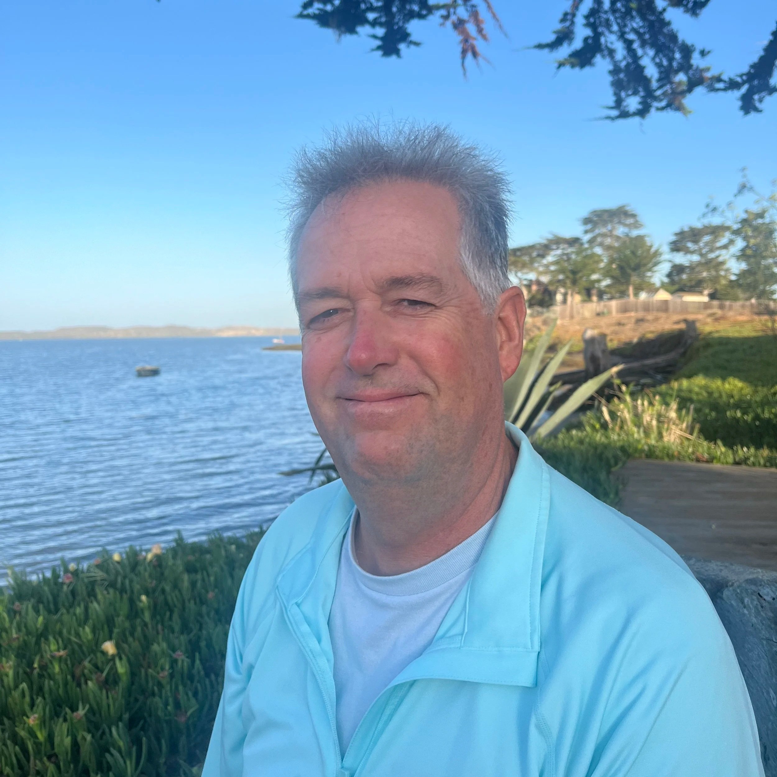 Close-up of a middle-aged man with short, gray hair, smiling, outdoors near a lake with trees and a clear blue sky in the background.