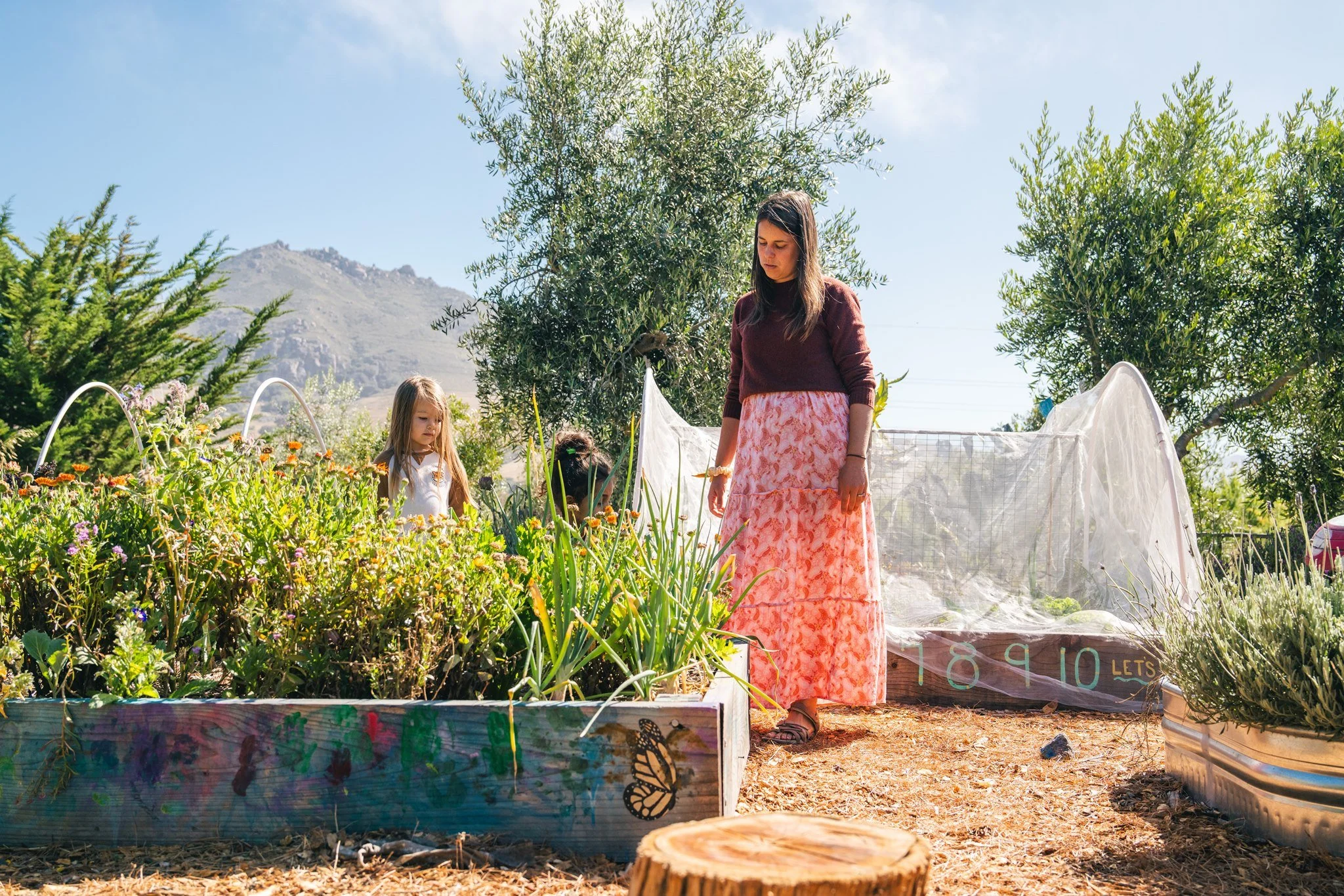 A woman and two young girls in a garden with flowering plants, trees, and mountains in the background. The woman is wearing a maroon sweater and a pink patterned skirt, while the girls are dressed in white. There are garden decorations and a clear blue sky.