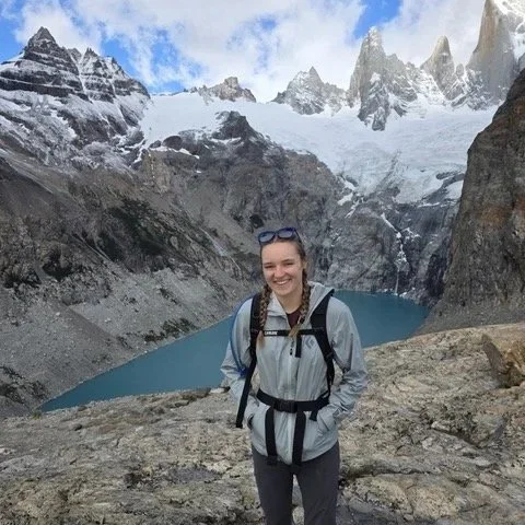 A woman hiking in the mountains with snow-capped peaks, a glacier, and a turquoise lake in the background.