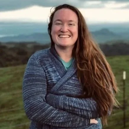 A woman with long red hair smiling outdoors in a grassy field with distant hills and cloudy sky in the background.