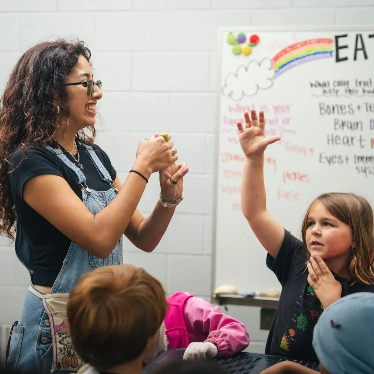 A woman with curly hair and glasses smiling and talking to a young girl raising her hand in a classroom. The girl has brown hair and is touching her neck. There are children sitting at a table and a whiteboard with colorful drawings and notes in the background.
