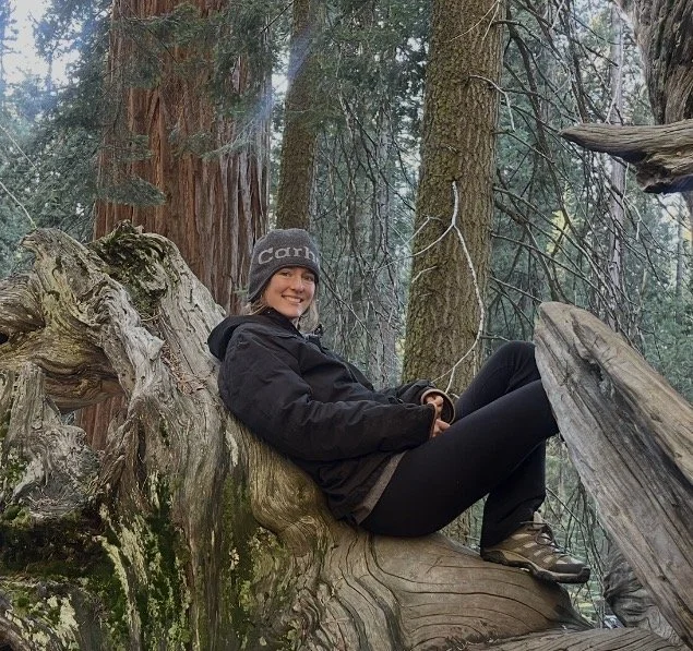A woman sitting on a large fallen tree trunk in a forest, smiling and wearing outdoor gear including a gray beanie, black jacket, and hiking boots.