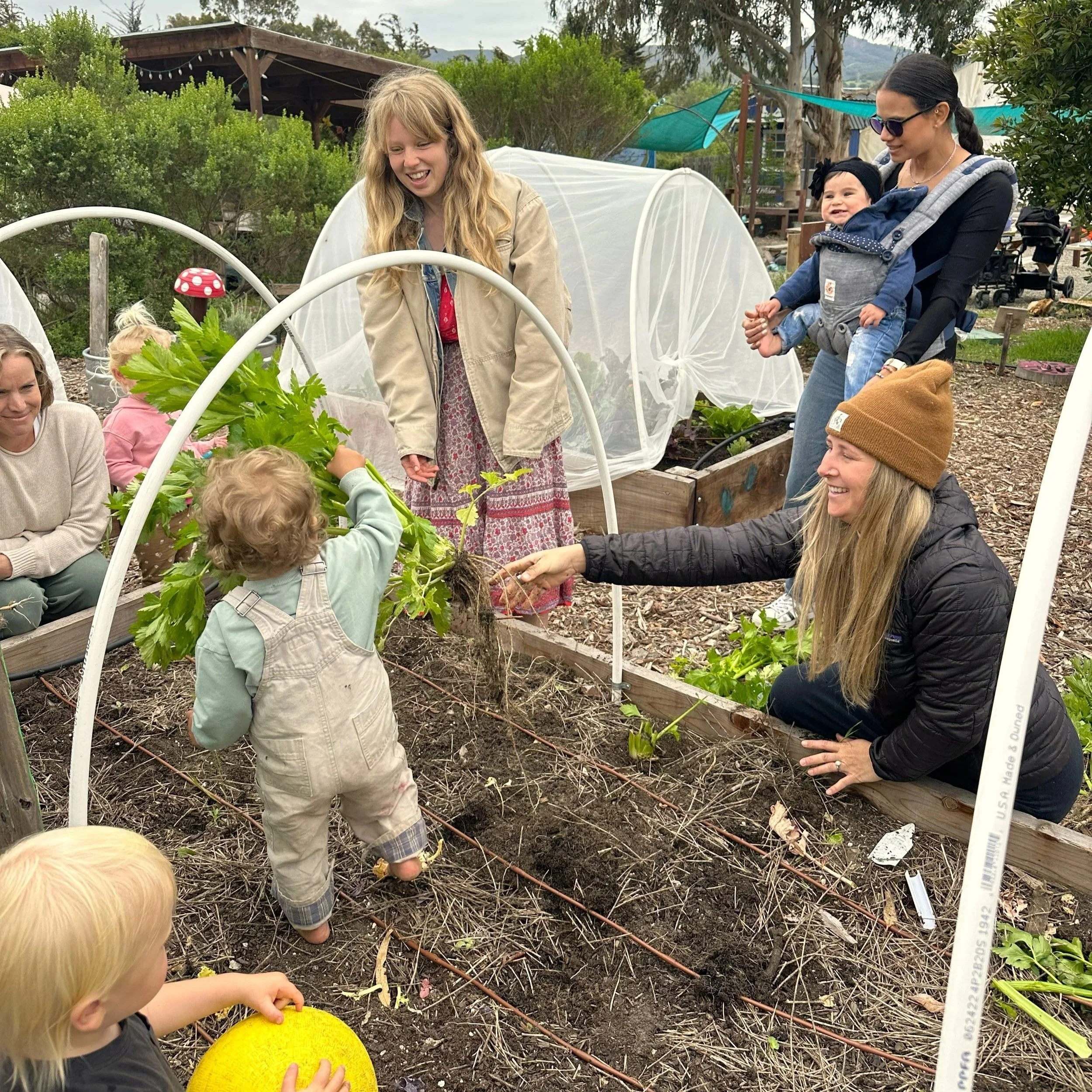 Children and adult in a garden with green plants, some children are reaching towards the plants while an adult is smiling and talking to them, and a woman is holding a child in a baby carrier, all surrounded by garden beds and garden structures.