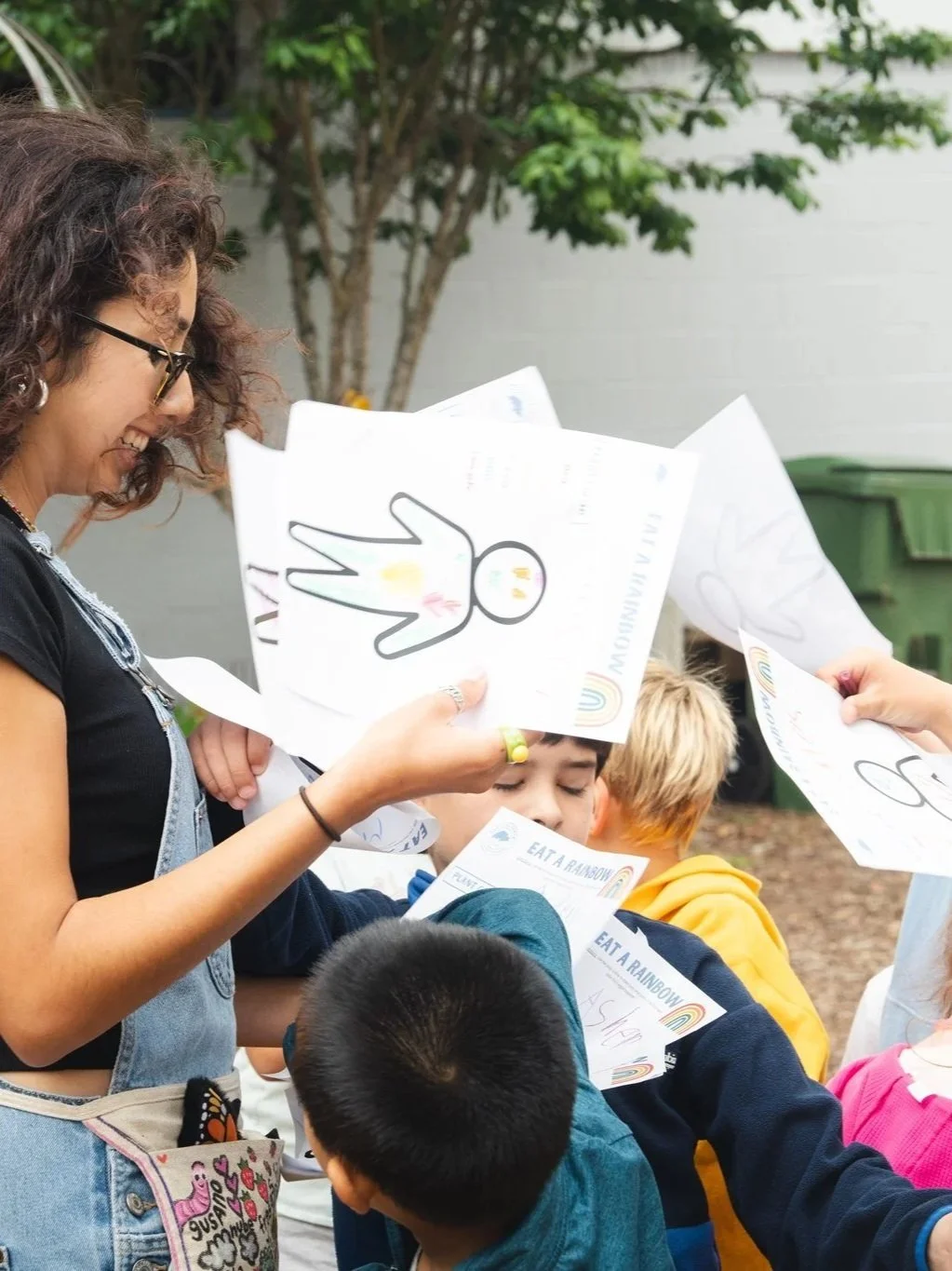 Children and a woman holding up colorful flyers or drawings outdoors, with trees and a building in the background.