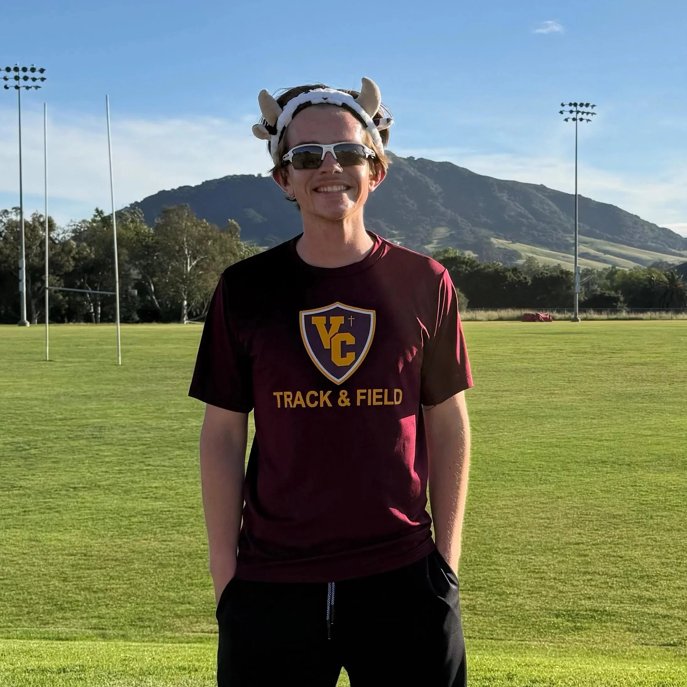 A young man smiling outdoors at a track and field stadium, wearing a maroon shirt with yellow lettering and a hat with horns and a headband, with mountains and a blue sky in the background.