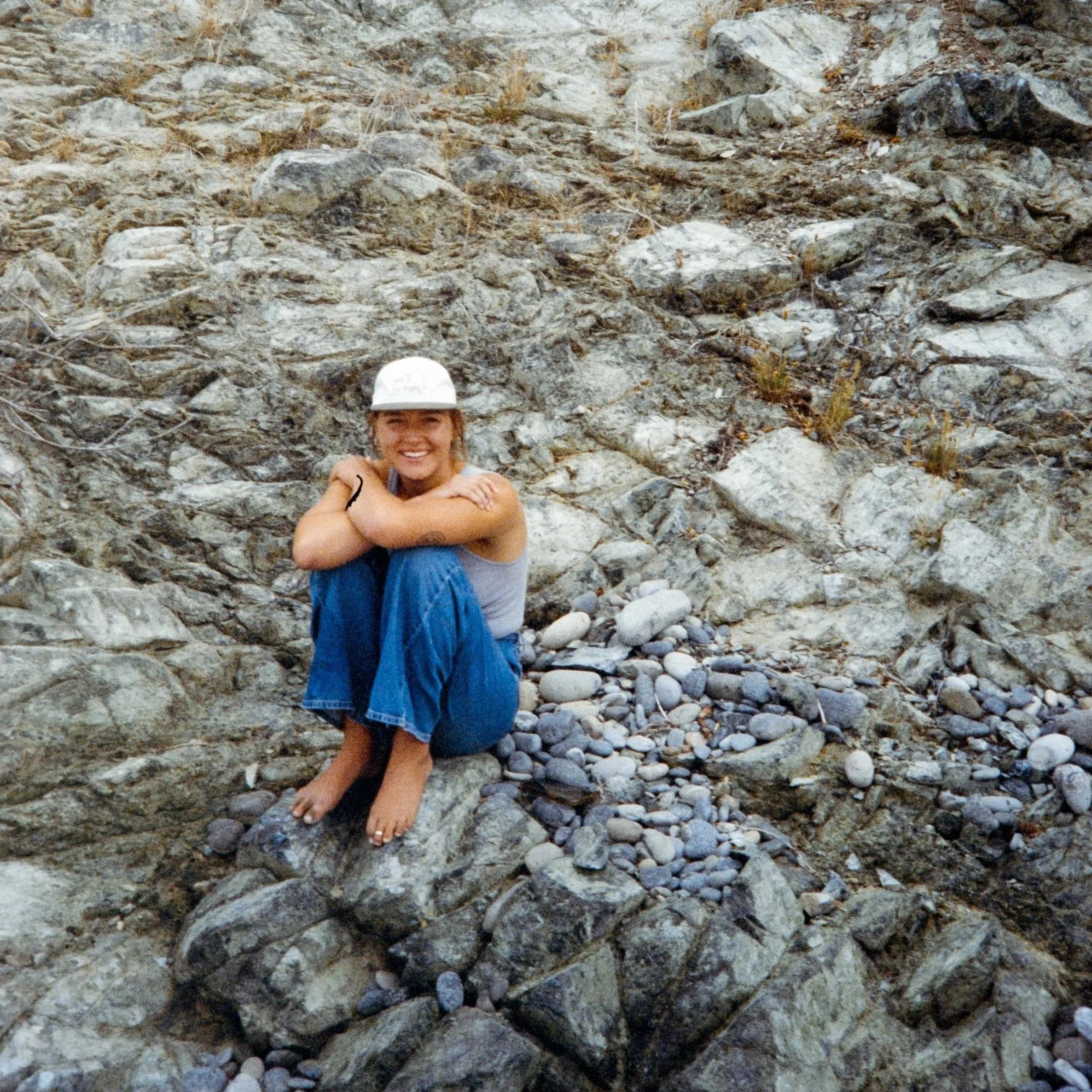 A woman with a white hat sitting on a rocky hillside with a broad smile.