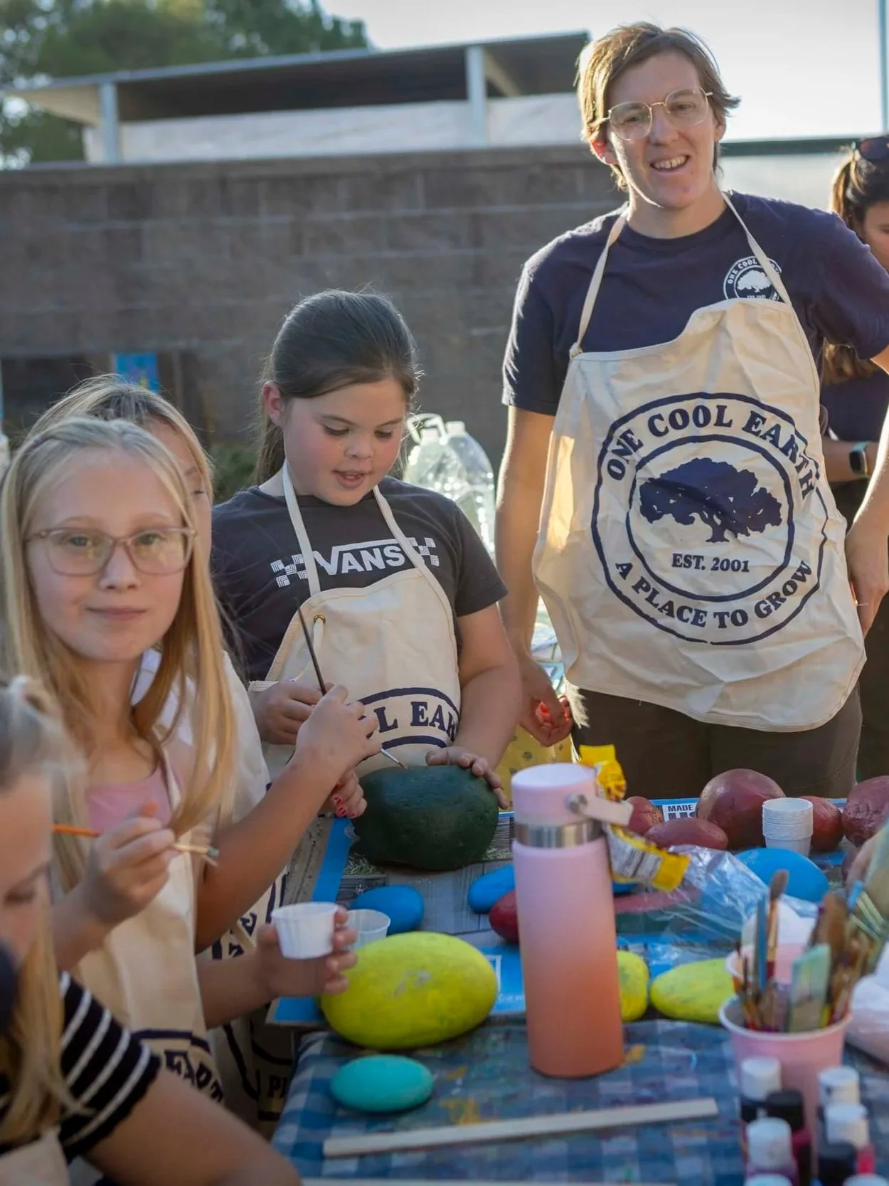 Children and adult decorating Easter eggs outdoors at a table with colorful eggs, cups, and craft supplies.
