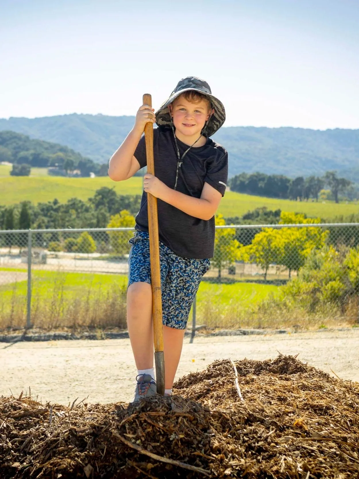 A young boy wearing a wide-brimmed hat, black shirt, and patterned shorts standing outdoors in a field with a shovel, planting or digging in the soil, with a backdrop of green hills and blue sky.