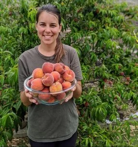 Woman smiling and holding a bowl of peaches outdoors in a garden.