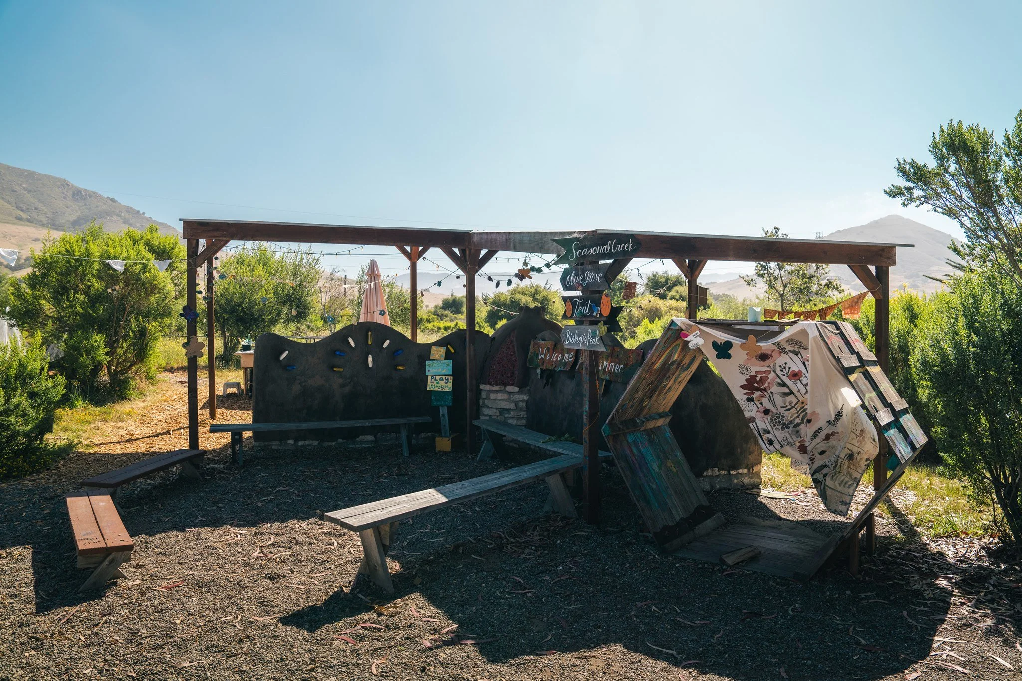 Wooden outdoor shelter with handmade signs, surrounded by greenery and mountain landscape under clear blue sky.