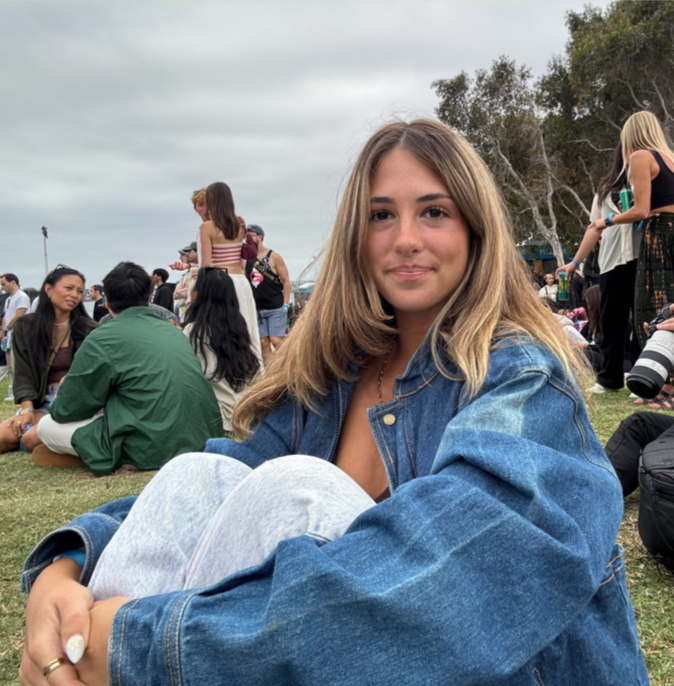A young woman with light brown hair, wearing a denim jacket and gray pants, sitting on the grass at an outdoor event with many people around, some sitting and some standing, under cloudy skies.