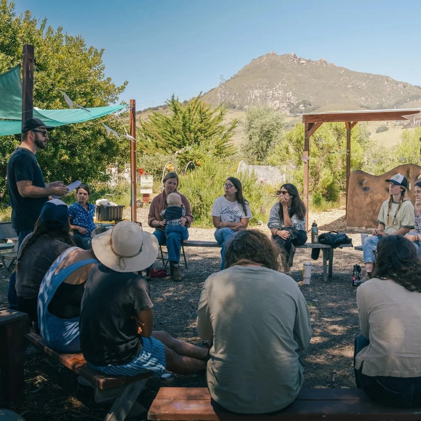 Group of people sitting outdoors on benches and listening to a man speaking, with a mountain and trees in the background.