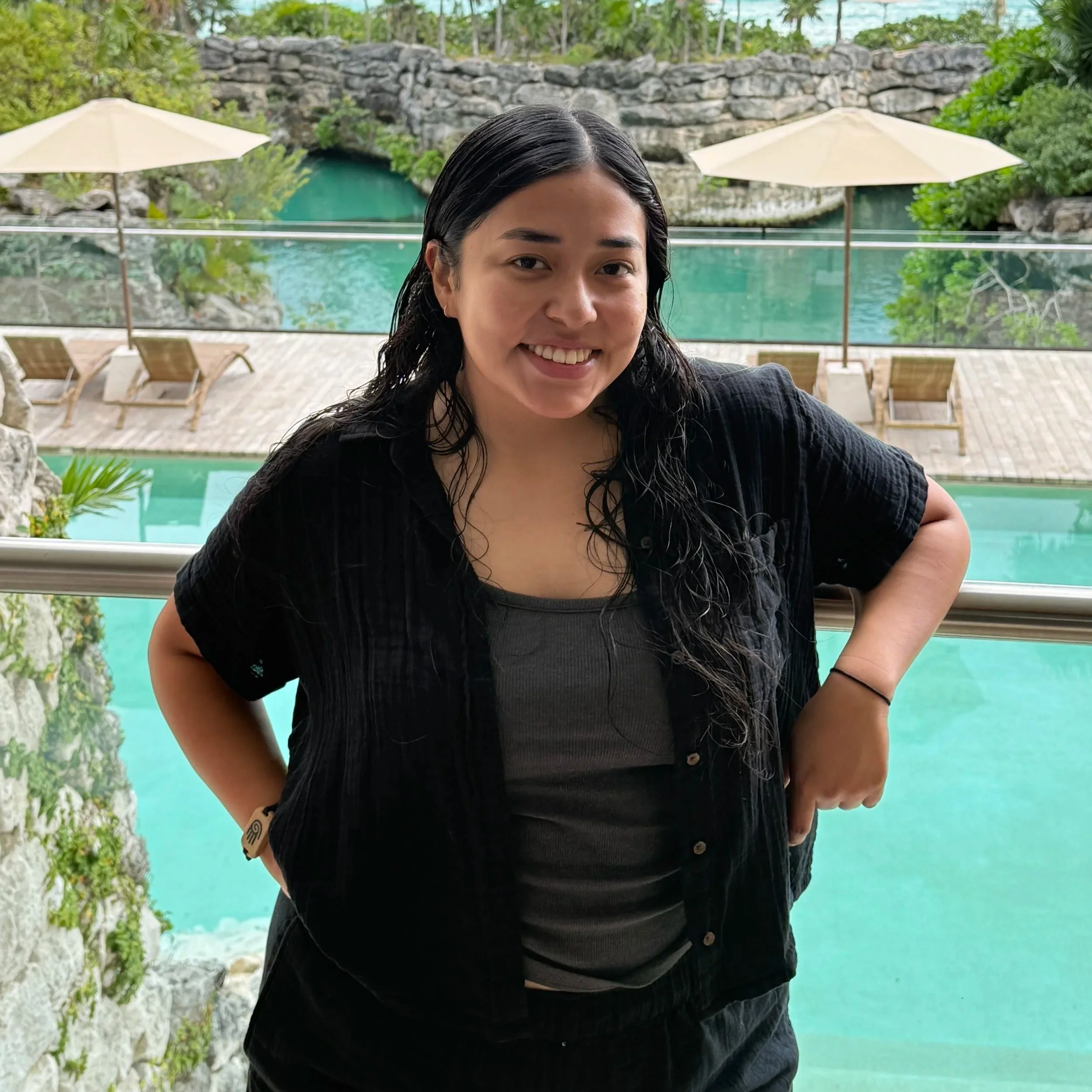 A smiling woman with long, dark hair standing outdoors near a pool with a rocky landscape and umbrellas in the background.