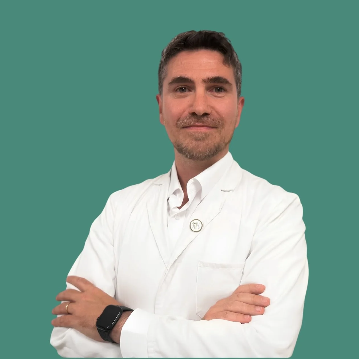 Hombre con barba, cabello castaño, usando bata blanca y reloj inteligente, con los brazos cruzados frente a fondo verde.