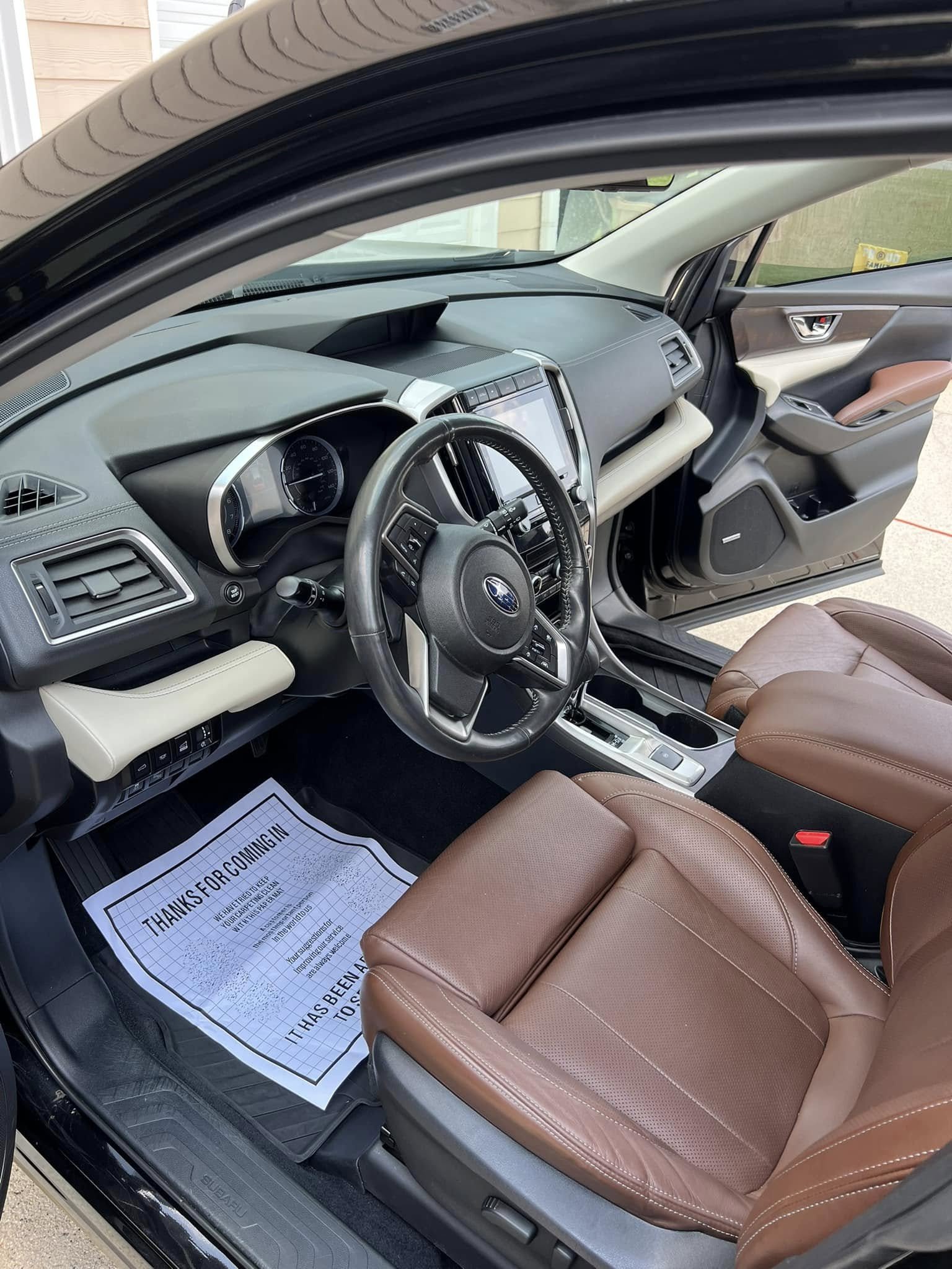 The interior of a modern vehicle showing the driver's seat, dashboard, steering wheel, and a paper floor mat with a message. The seats are tan leather and the dashboard is black with a large center touchscreen.