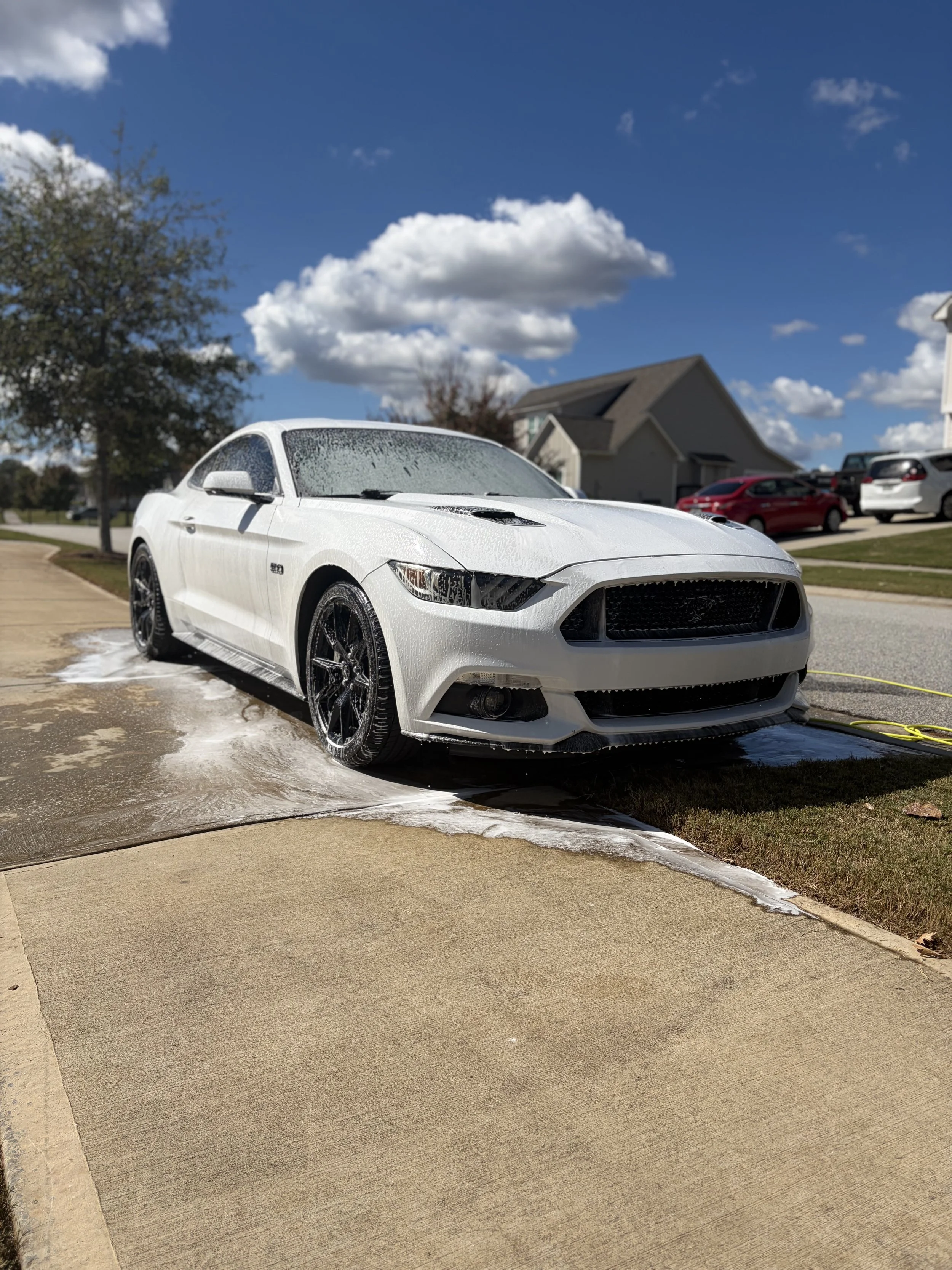 White sports car being washed with soap, parked on a driveway in a suburban neighborhood under a partly cloudy sky.