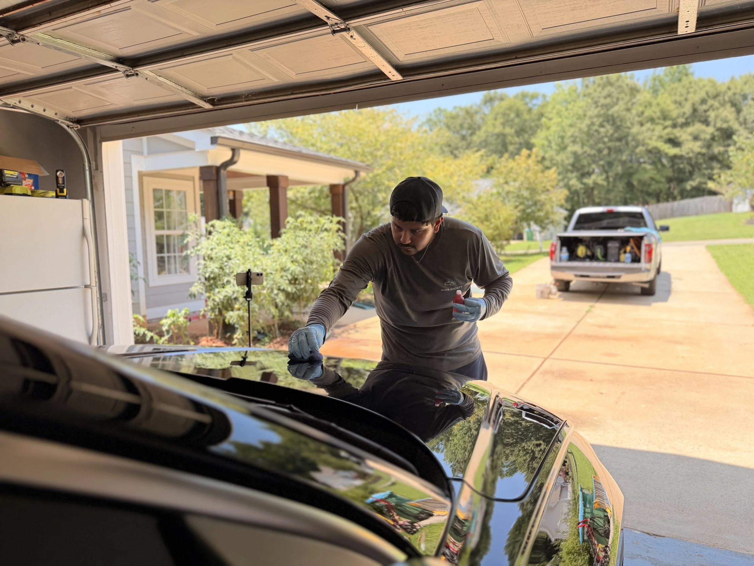 A person wearing a black cap and gloves working on a car in a garage with a view of green trees and a truck outside.